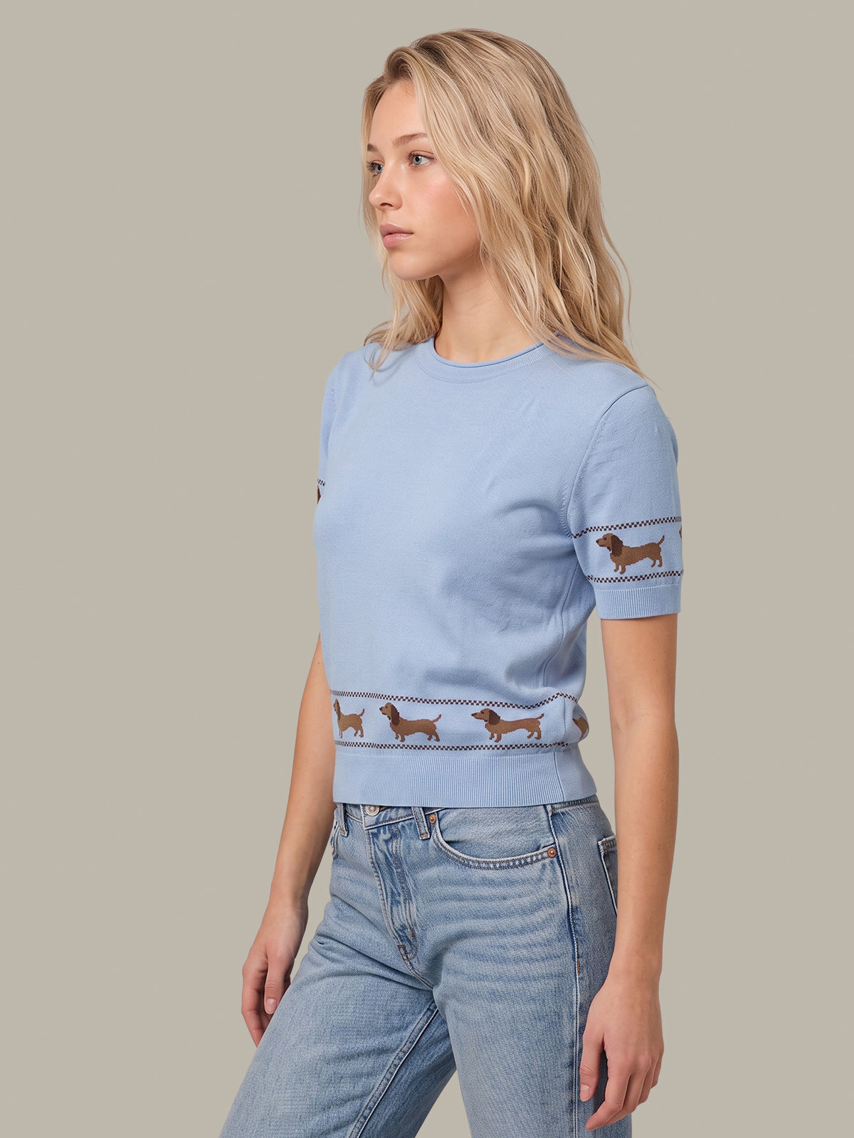 A woman wearing a light blue crewneck knit sweater with brown dachshund dog motifs along the hem and sleeve cuffs, paired with light-wash blue jeans, set against a solid taupe background.