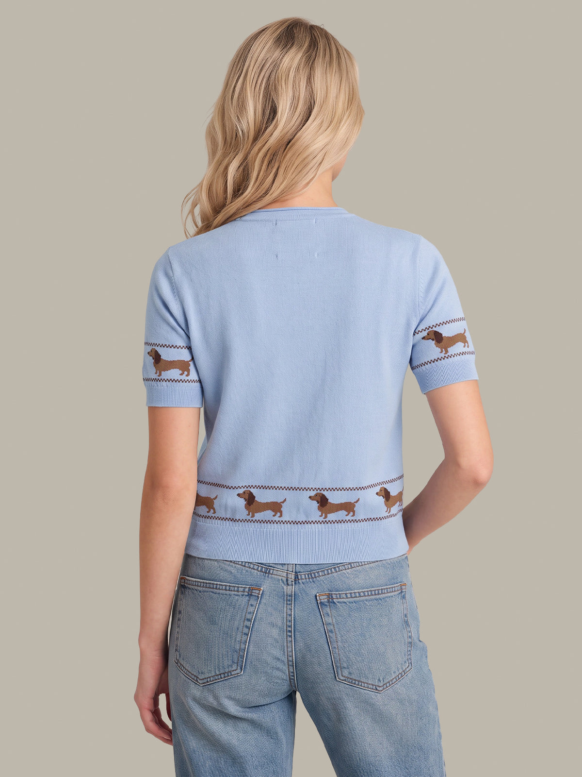 A woman shown from the back wearing a light blue short-sleeve knit top with brown dachshund dog embroidery across the shoulders, cuffs, and lower back, paired with light-wash jeans against a solid beige background.