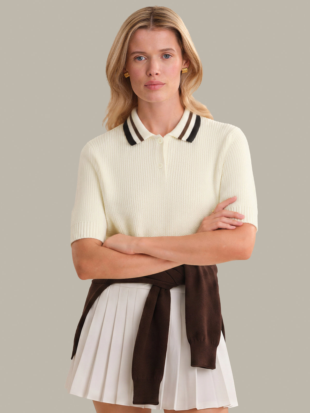 Woman wearing a cream-colored polo shirt with black trim and a white pleated skirt on a gray background
