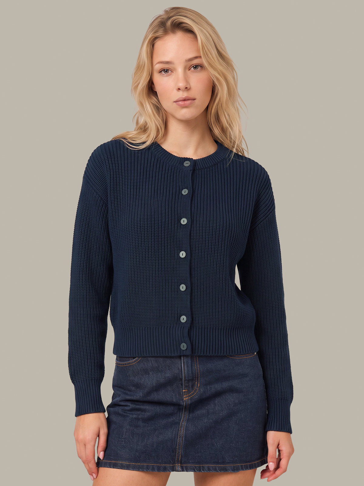 A blonde woman modeling a dark navy ribbed knit cardigan with a button-up front, paired with a dark denim mini skirt, against a solid light gray background.