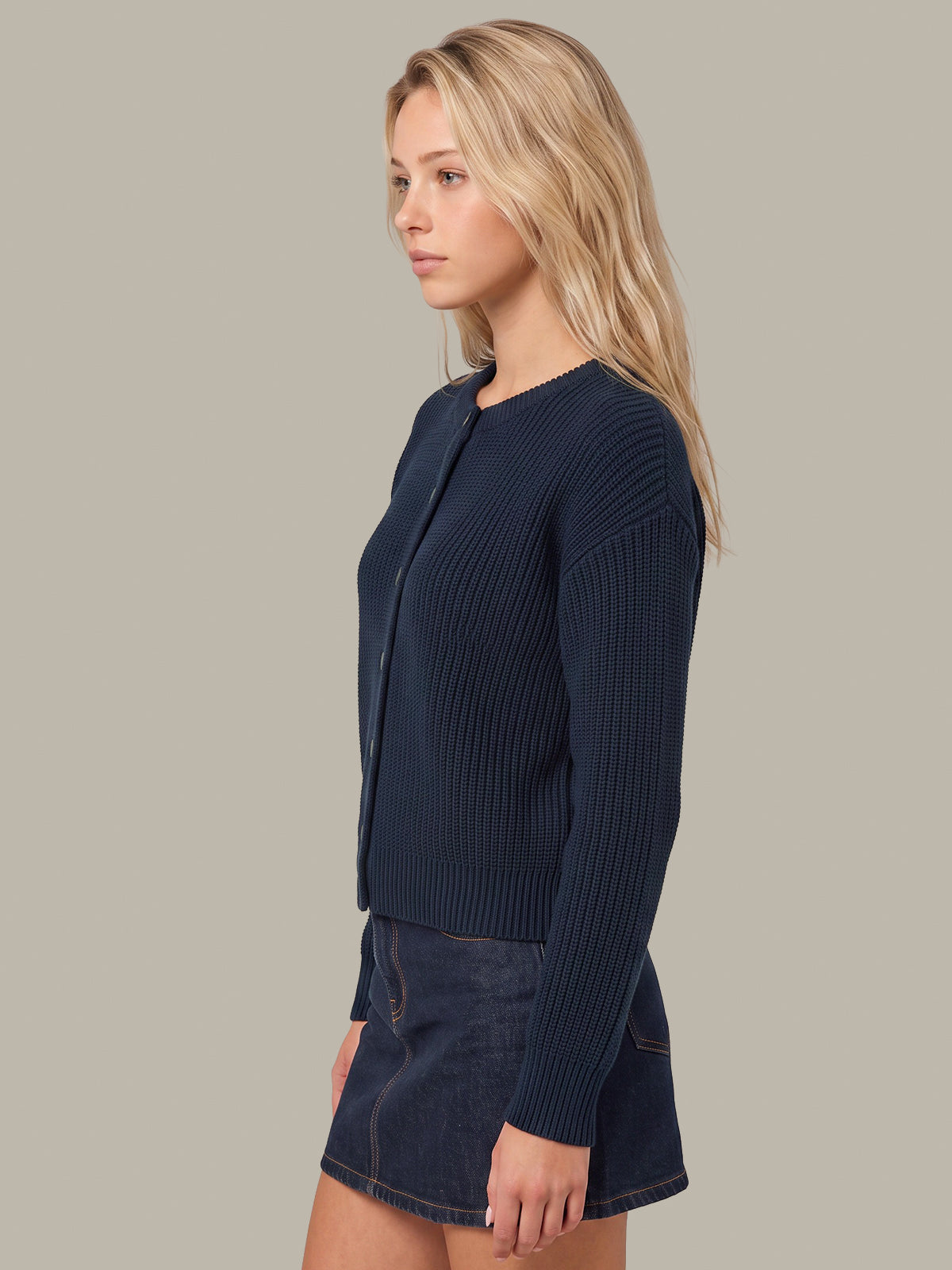 A woman shown in side profile wearing a dark navy ribbed knit cardigan and a dark denim mini skirt against a solid light gray studio background.