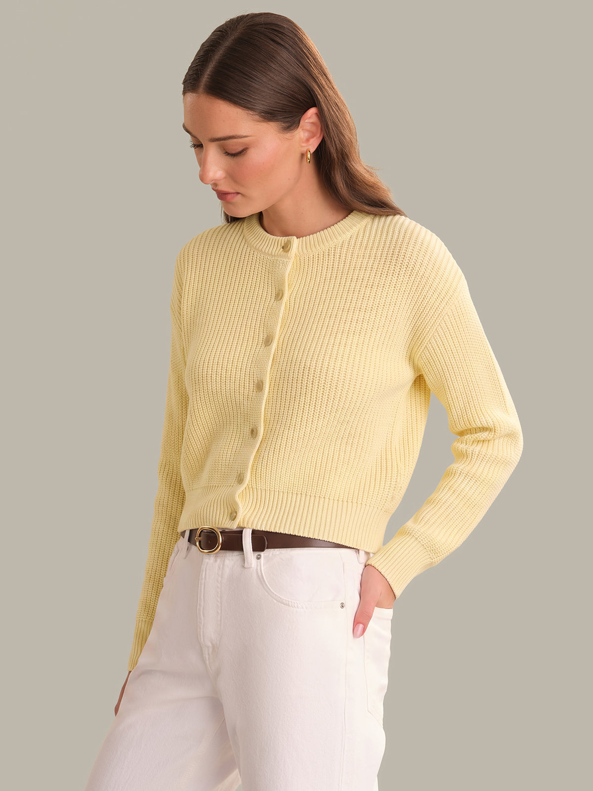 Woman wearing a light yellow ribbed cardigan and white pants on a gray background
