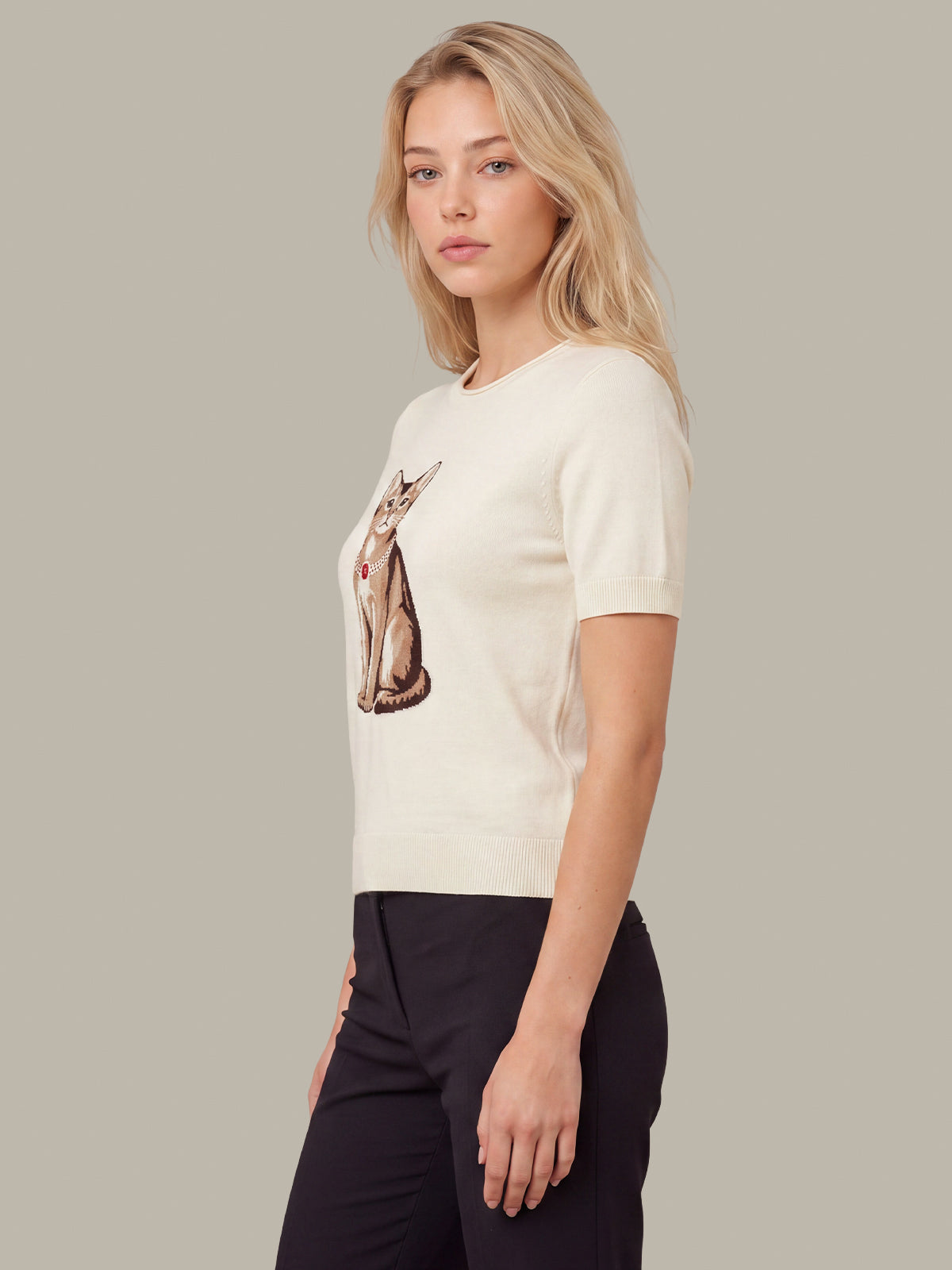 A blonde woman wearing a cream short-sleeve knit top with a detailed cat graphic on the front, paired with black pants, set against a solid gray studio background.