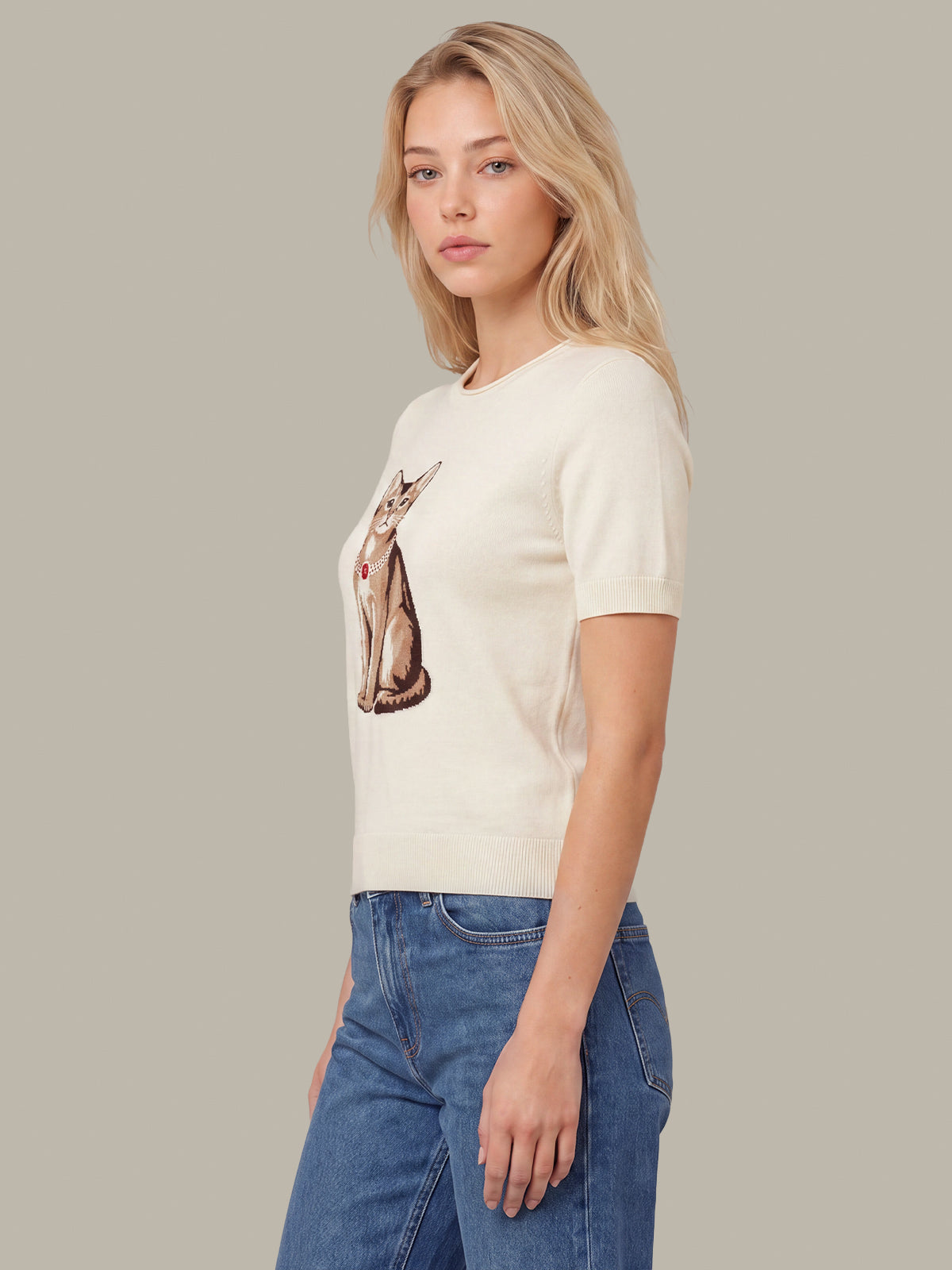 A blonde woman wearing a cream short-sleeve crewneck t-shirt with a detailed cat graphic on the front, paired with blue jeans, set against a solid taupe background.