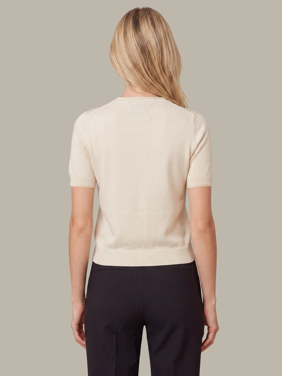 A woman viewed from the back wearing a short-sleeve cream knit top and dark trousers, against a plain warm gray studio background.