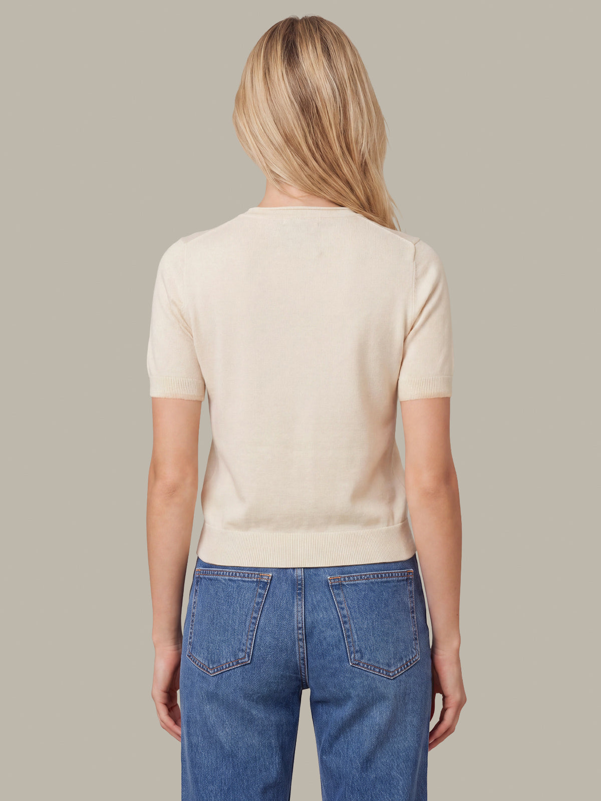 A woman viewed from the back wearing a short-sleeve cream knit top and blue jeans against a solid taupe background.