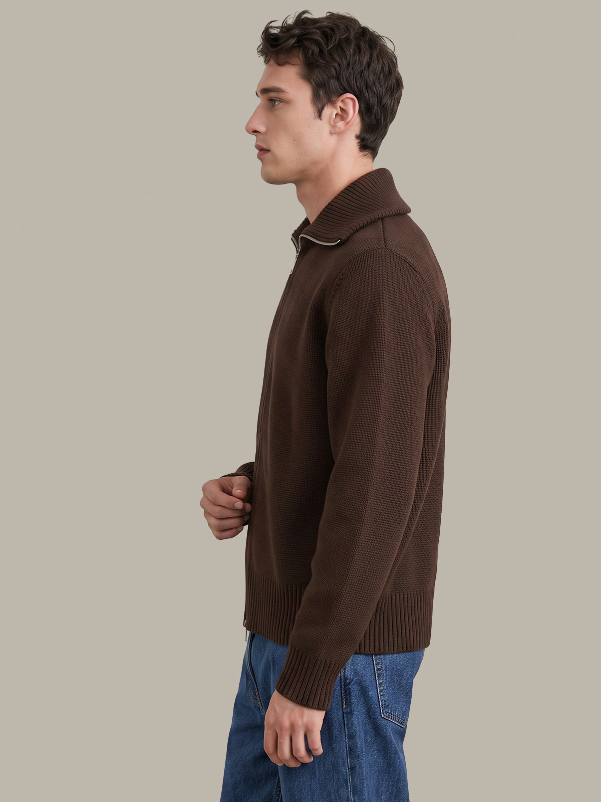 A man in side profile wearing a dark brown ribbed knit sweater with a high collar and blue jeans, set against a solid light gray background.