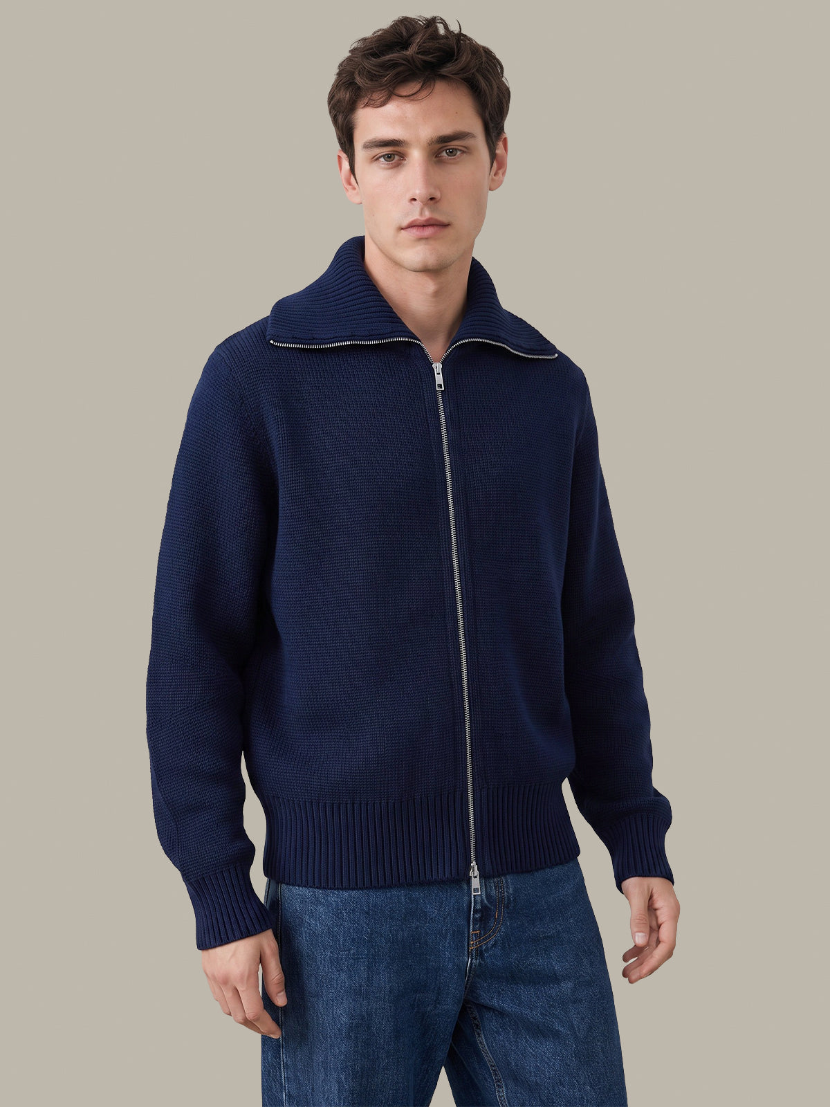 A man wearing a navy blue knit zip-up sweater with a ribbed mock neck, cuffs, and hem, paired with blue jeans, against a solid light gray background.