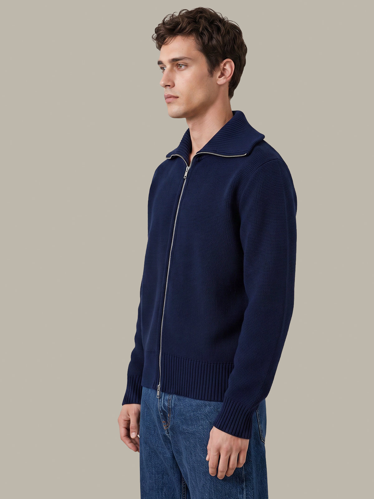 A man wearing a navy blue zip-up knit sweater with a stand collar and light piping detail, paired with blue jeans, photographed against a solid light gray background.