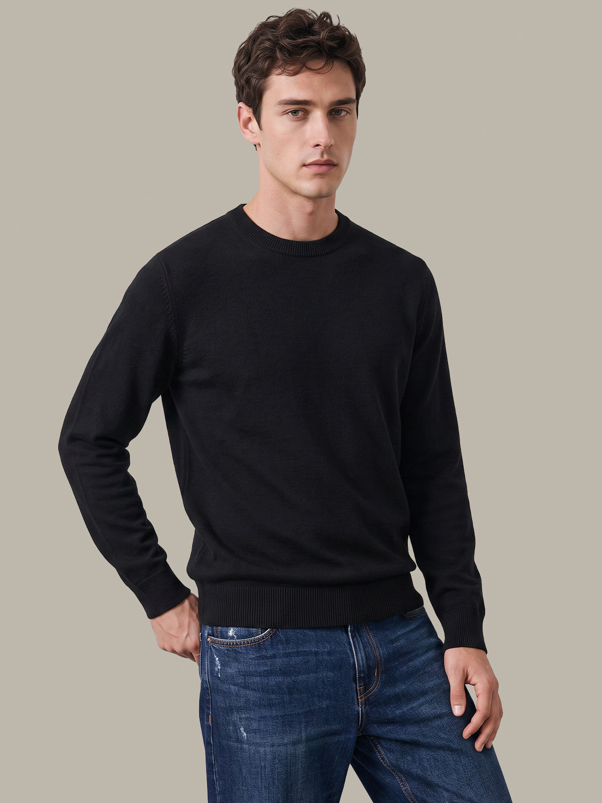 A man wearing a black long-sleeve crewneck sweater and blue jeans, standing against a solid light gray background.