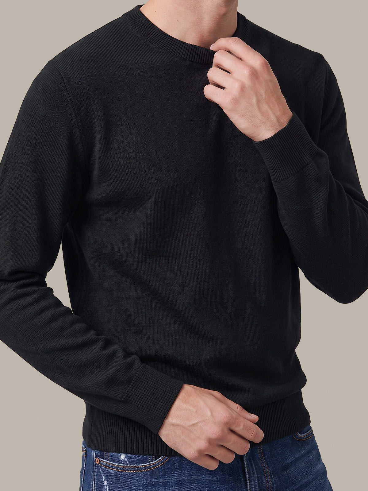 A man wearing a black long-sleeve crewneck sweater, paired with blue jeans, shown from the neck down against a solid neutral background.