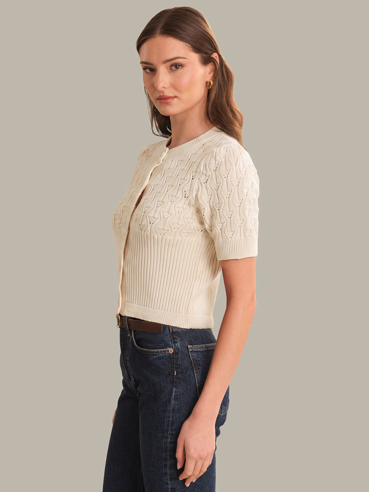 Woman wearing a cream knit cardigan over a ribbed top with dark jeans on a gray background