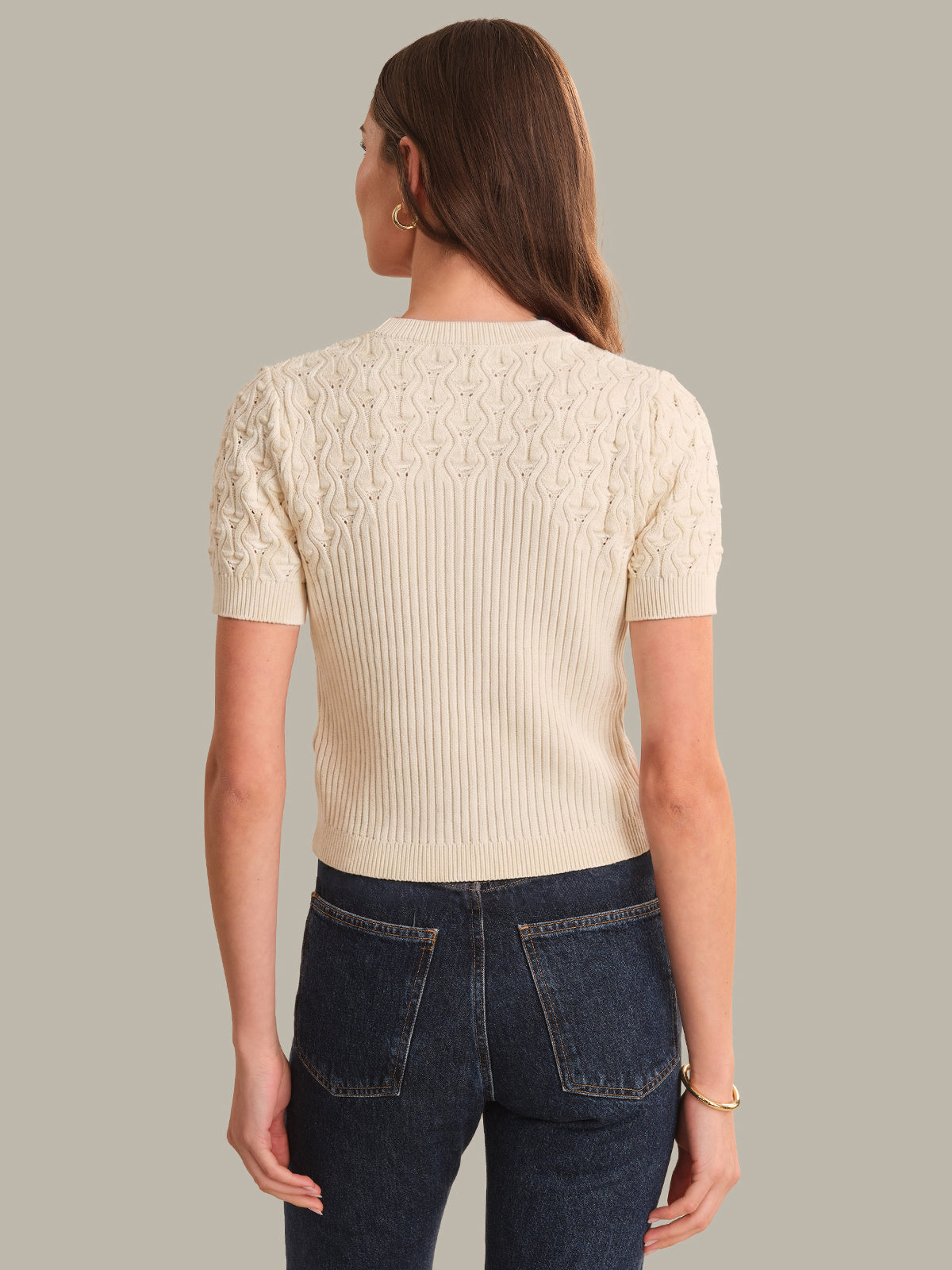 Woman wearing a cream ribbed cardigan and dark blue jeans on a gray background