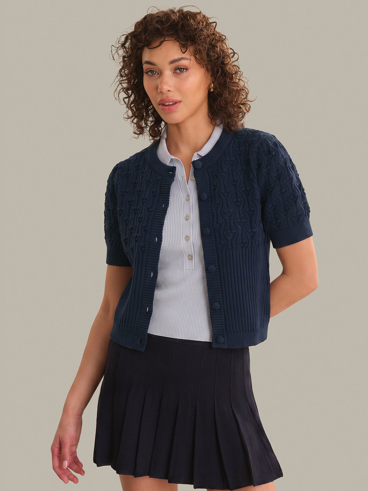 Woman wearing a navy blue cardigan over a white shirt and black pleated skirt on a gray background