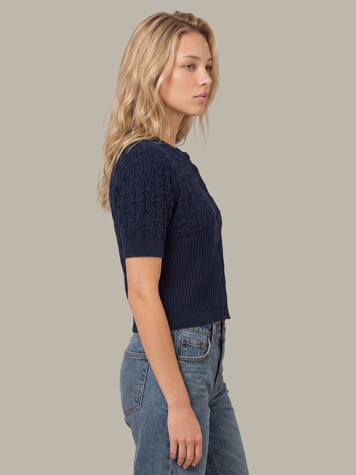 A woman in side profile wearing a navy blue short-sleeve knit top with textured geometric patterns on the upper chest and sleeves, paired with light-wash blue jeans, against a solid light gray background.