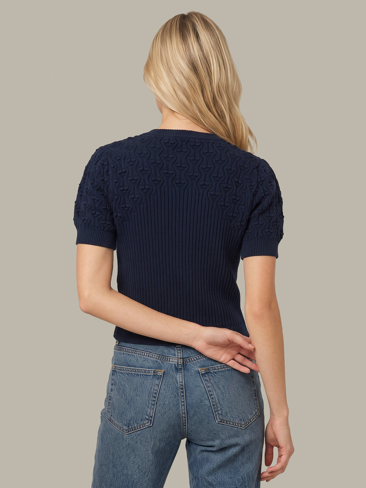A woman viewed from the back wearing a navy blue short-sleeve ribbed knit top with textured cable detailing on the shoulders and sleeves, paired with blue jeans, against a solid light gray background.