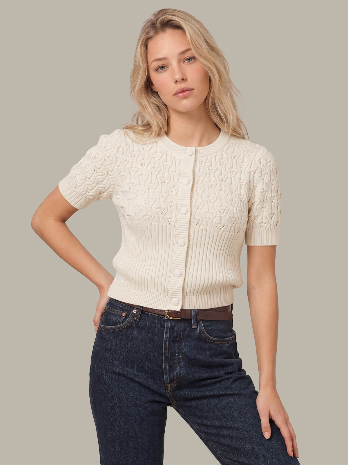 A blonde woman modeling a cream short-sleeve button-front cardigan with textured cable knit details, paired with dark blue jeans, against a solid light gray background.