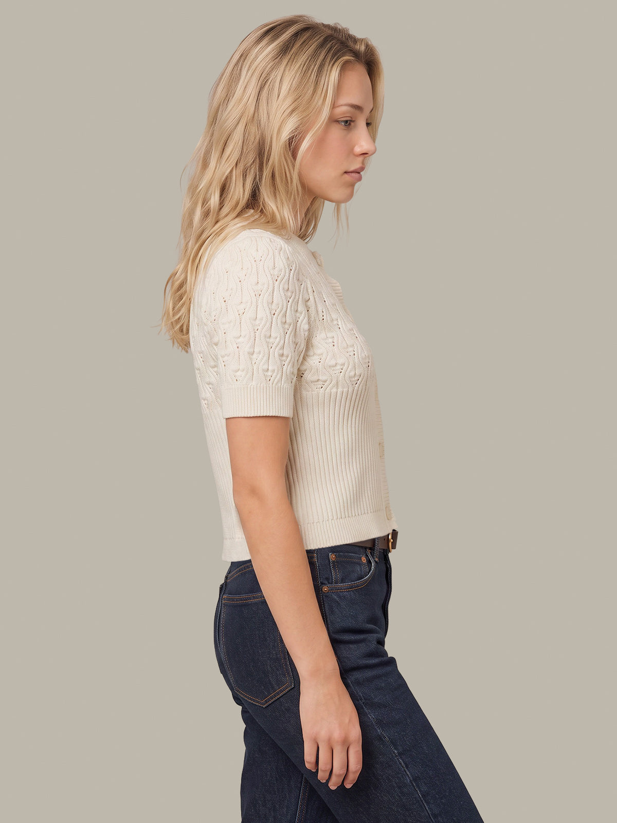 A woman in profile wearing a short-sleeve cream cable-knit top and dark blue jeans, photographed against a solid light gray studio background.