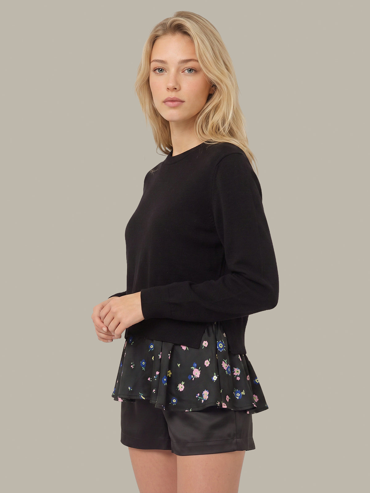 A blonde woman modeling a black long-sleeve crewneck top with a black peplum layer featuring a small multicolor floral print, paired with black shorts, against a solid taupe background.