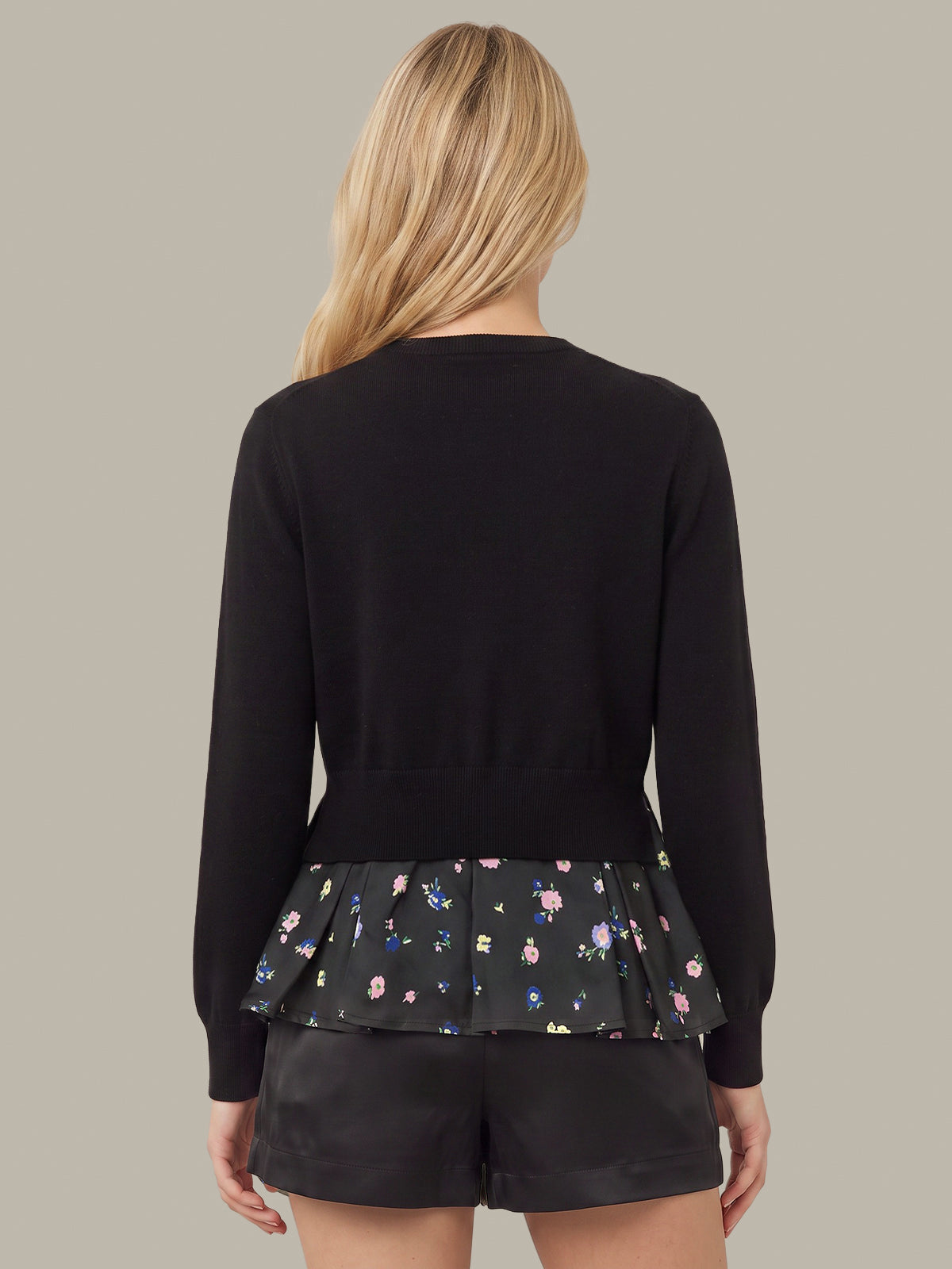 A woman shown from the back wearing a black long-sleeve cardigan with a layered ruffle hem featuring a small multicolor floral print, paired with black shorts, against a solid taupe background.