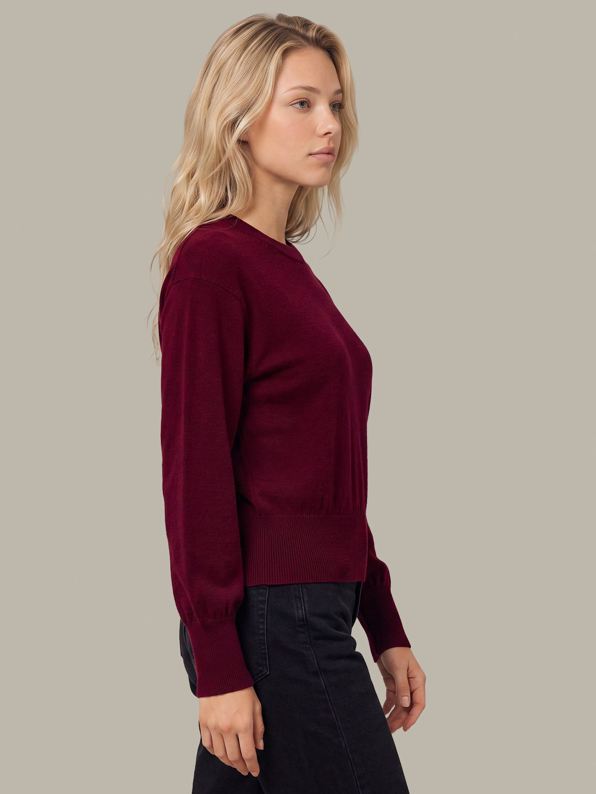A woman in profile wearing a long-sleeve crewneck sweater in deep burgundy, paired with dark jeans, against a solid light gray background.
