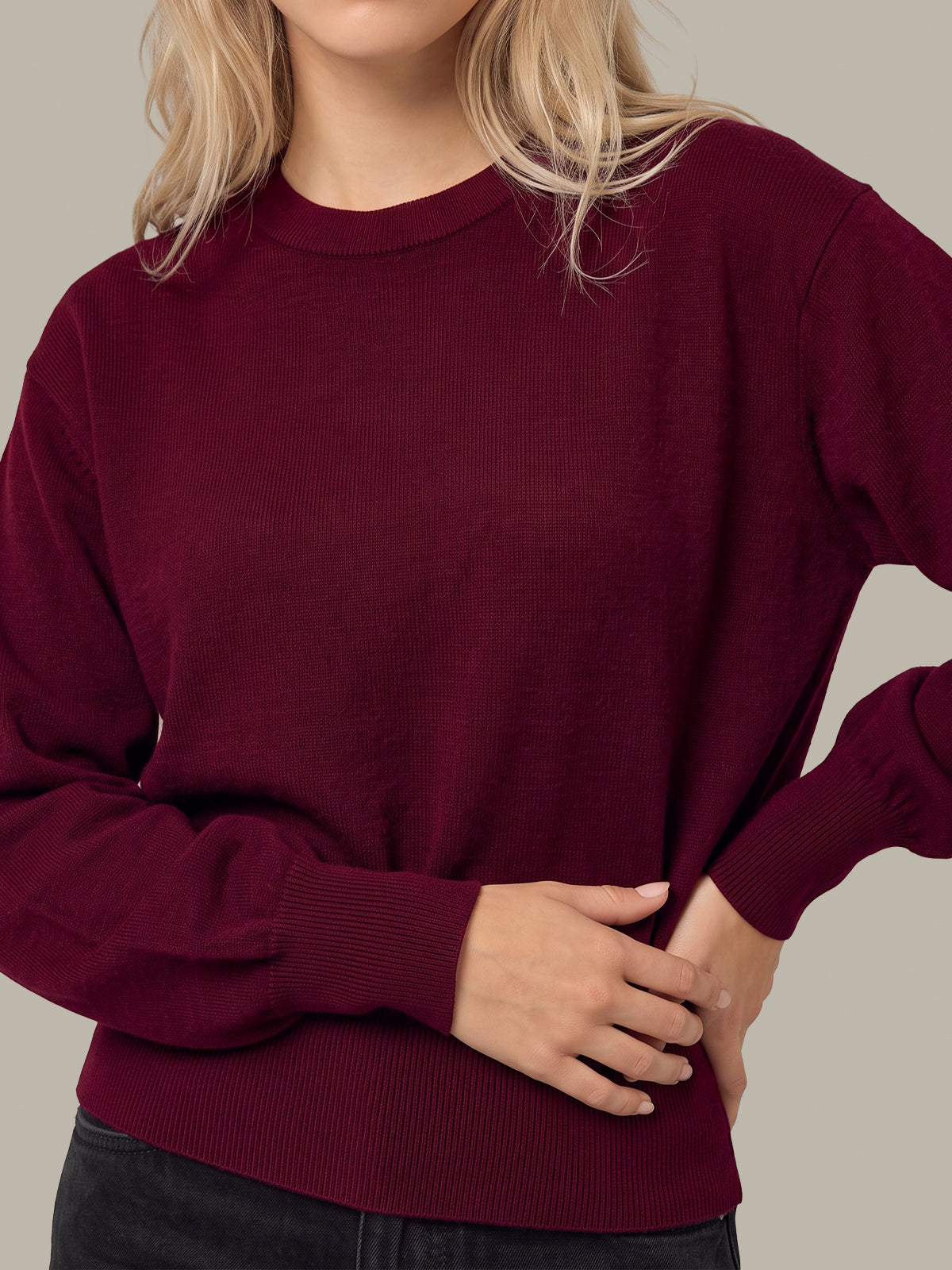 A woman wearing a long-sleeve crewneck sweater in deep burgundy, paired with dark jeans, against a solid light gray background.