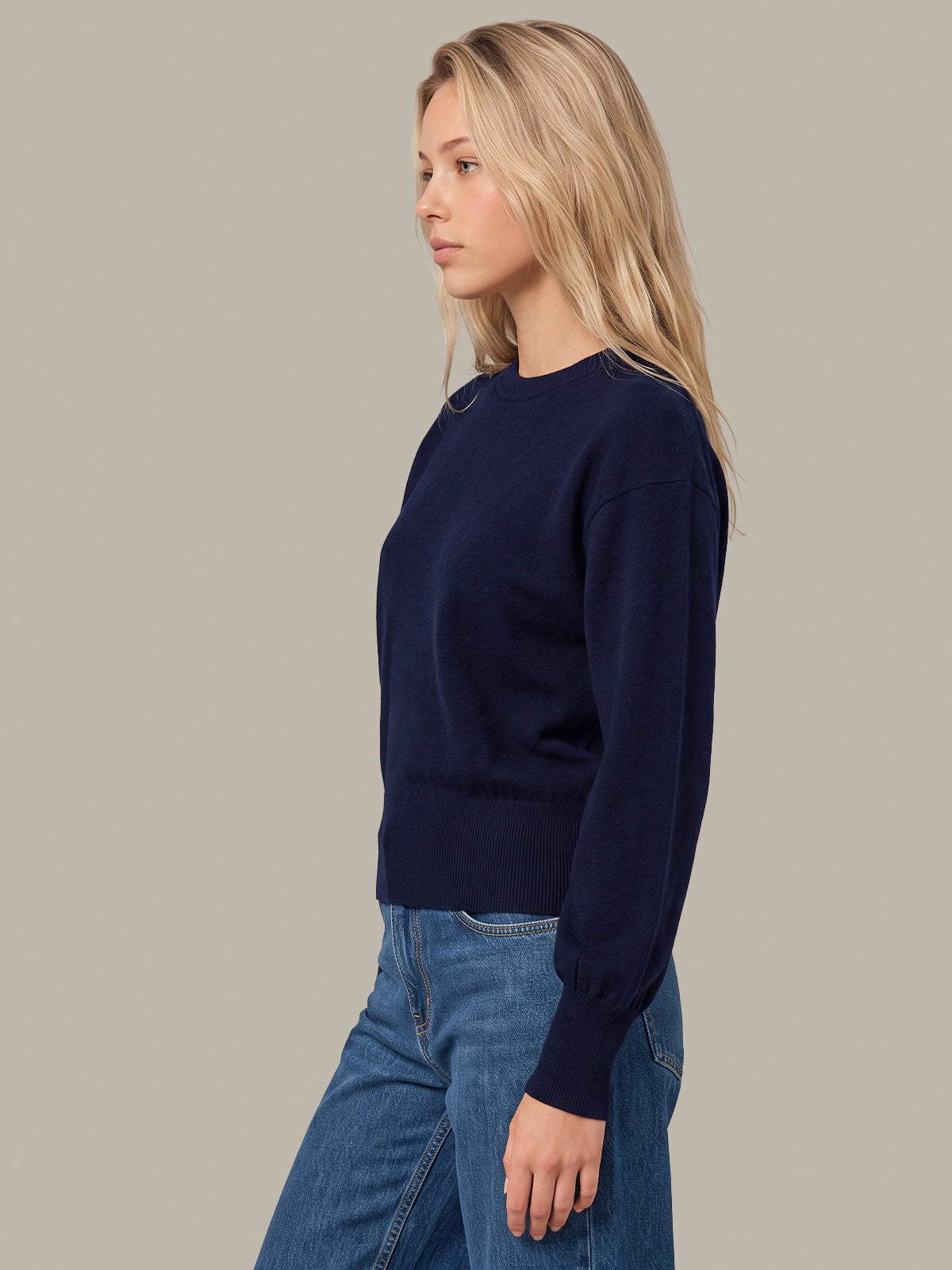 A woman in profile wearing a dark navy blue crewneck sweater and blue jeans, standing against a solid light gray background.