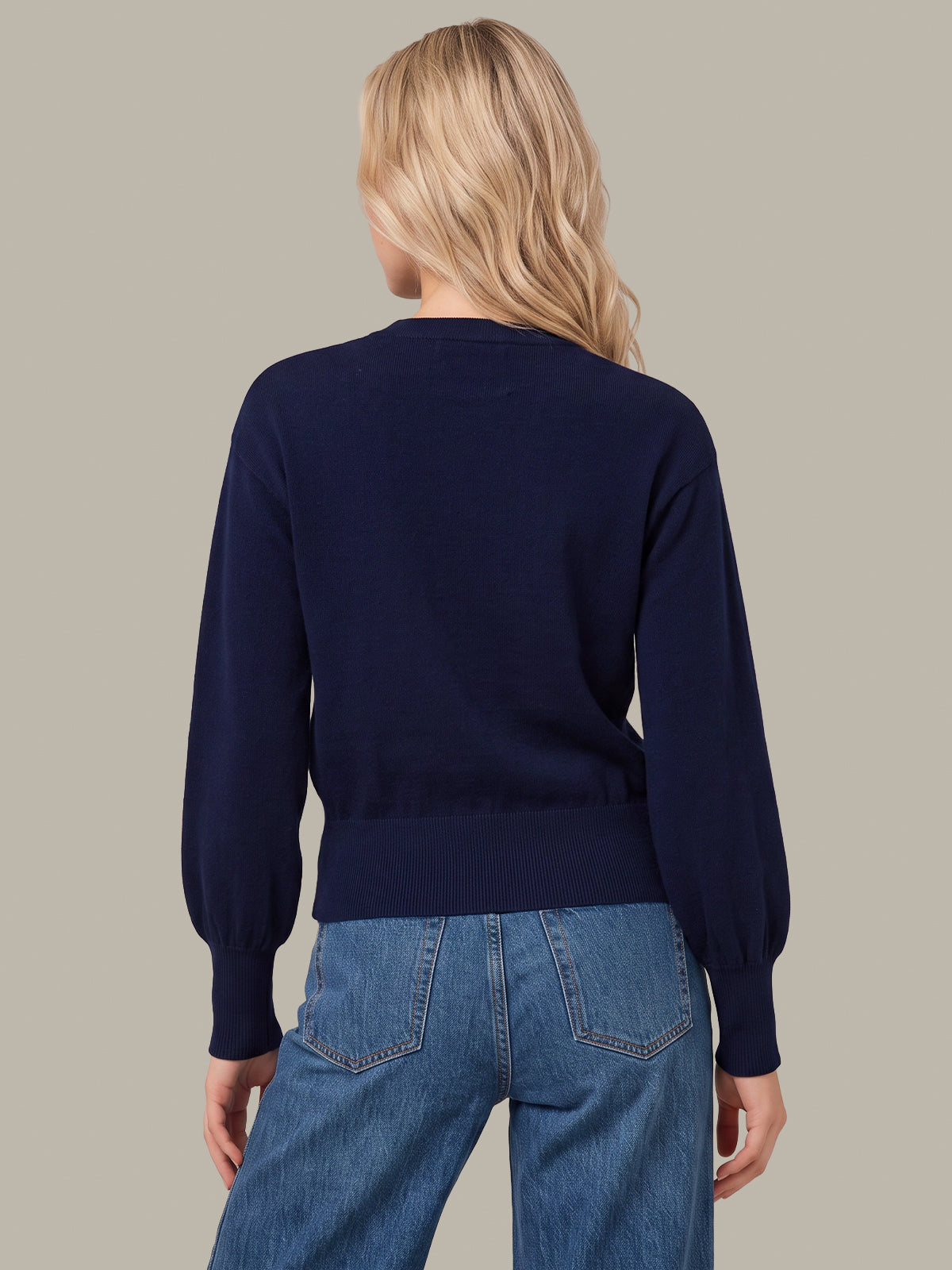 A woman viewed from the back wearing a navy blue long-sleeve knit sweater and blue jeans against a solid taupe background.