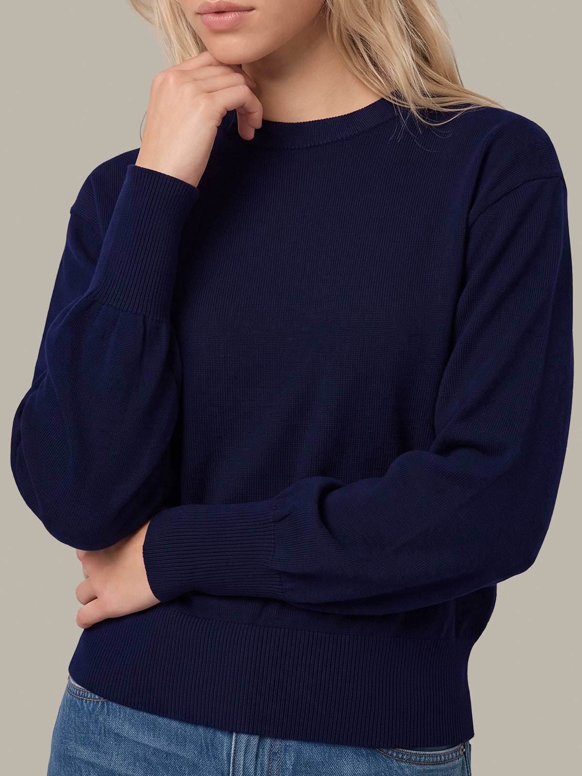 A woman wearing a navy blue long-sleeve knit sweater with ribbed cuffs, collar, and hem, paired with blue jeans, against a solid neutral background.