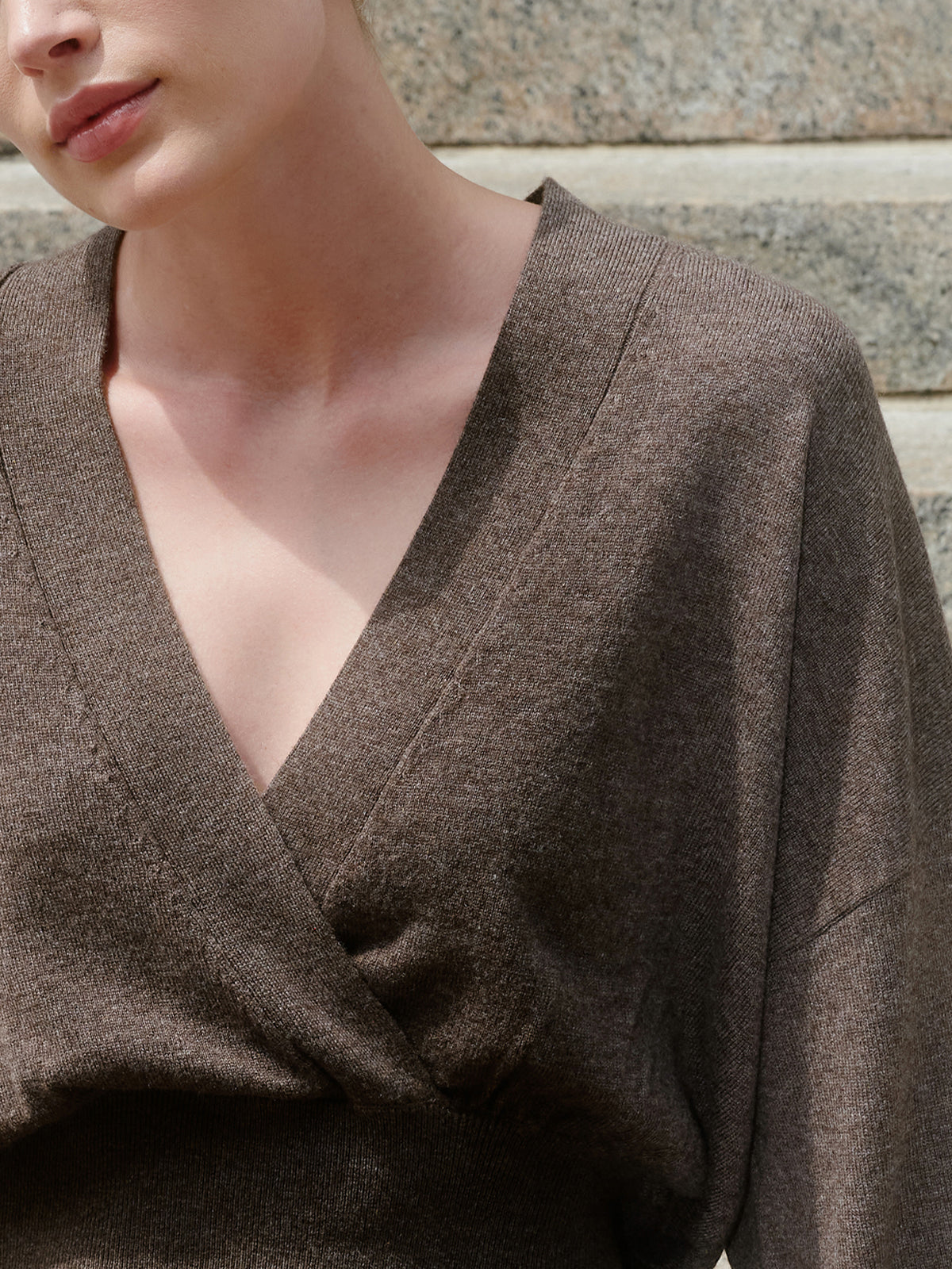 Close-up of a model wearing a dark brown textured knit wrap top with a deep V-neck, set against a neutral stone wall background.