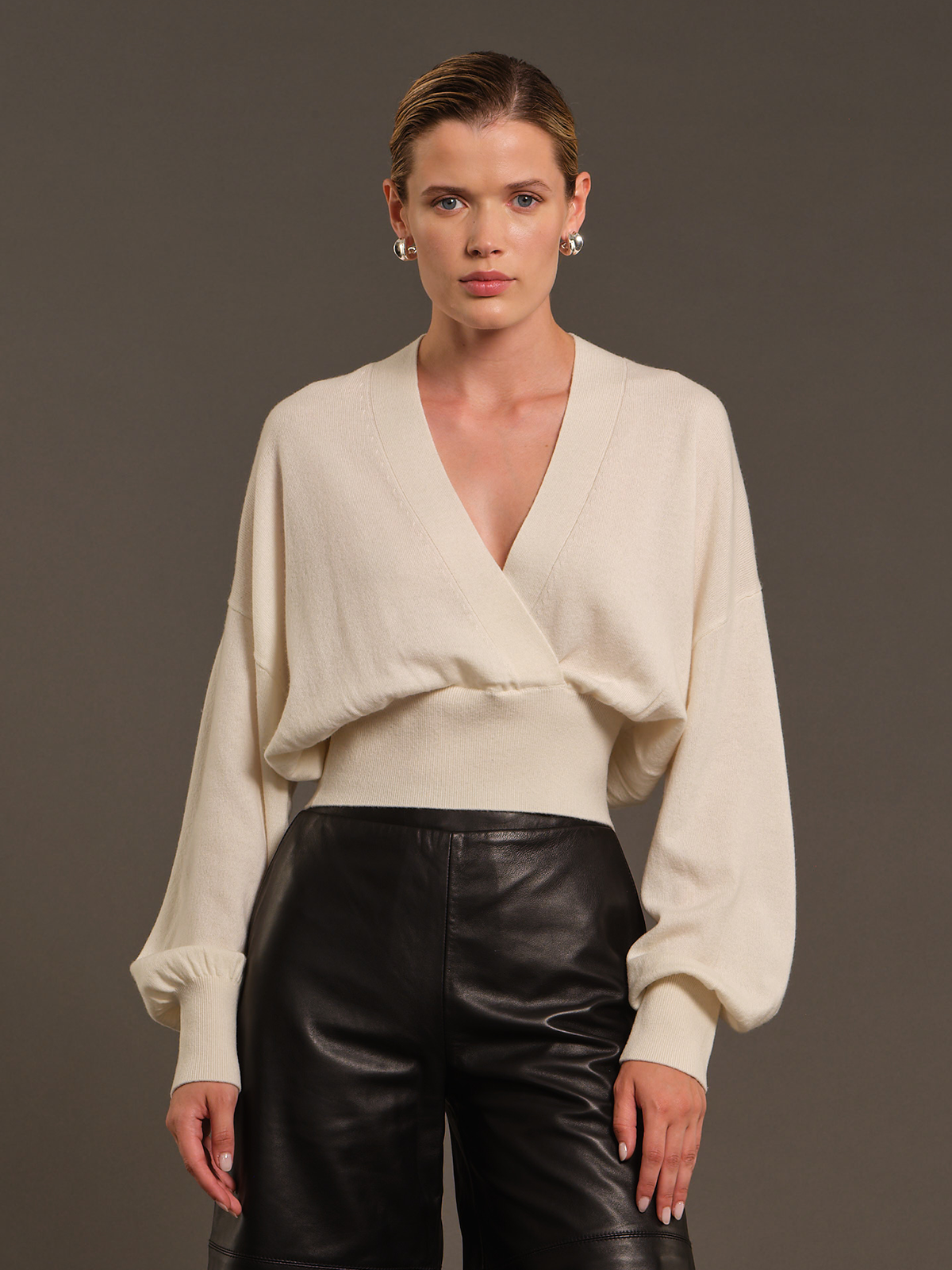 A model wearing a cream-colored wrap-style knit sweater with balloon sleeves and black leather pants, standing against a solid dark gray studio background.