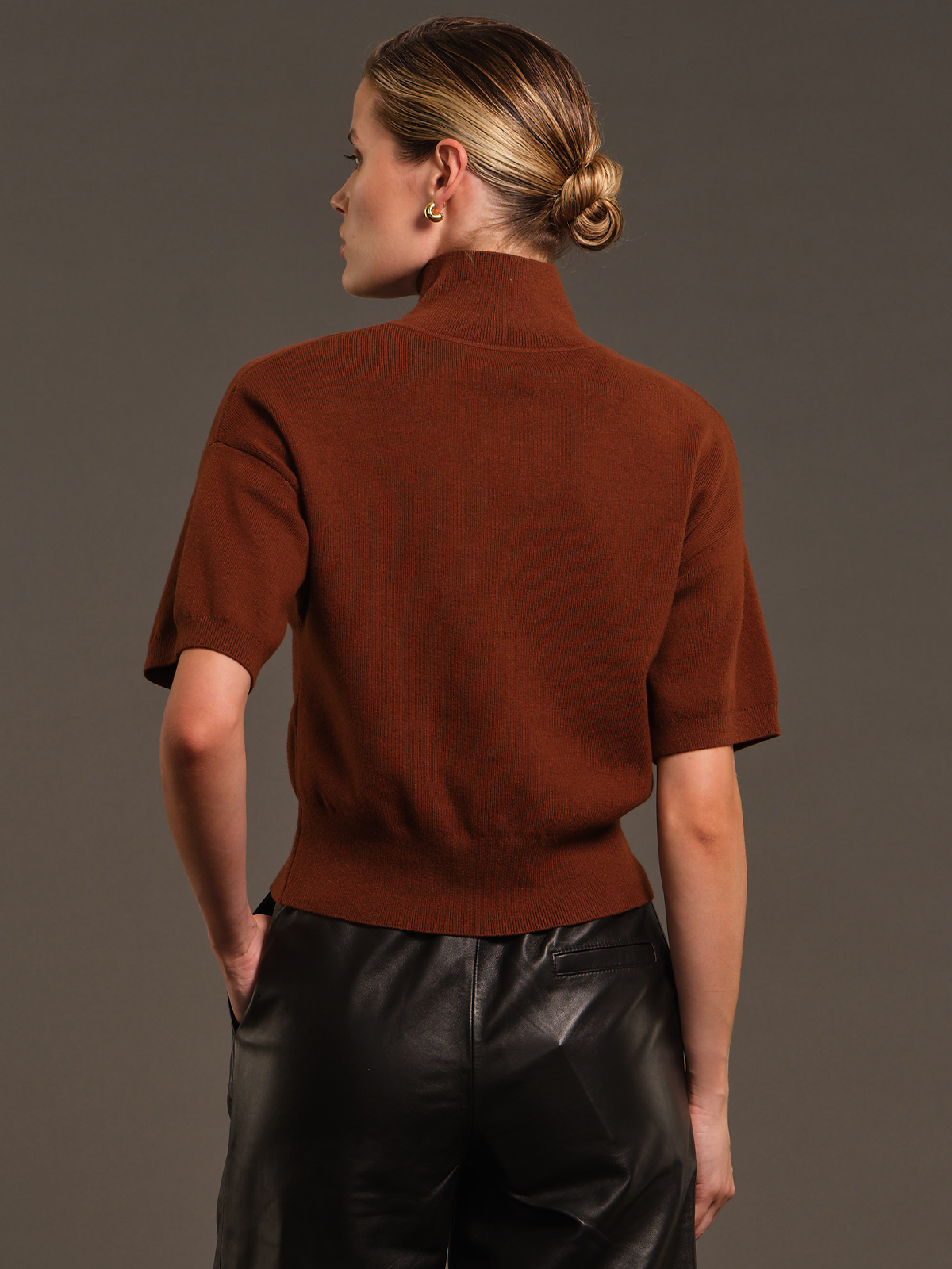 A woman shown from the back wearing a short-sleeve ribbed knit mock neck sweater in a warm rust brown color, paired with black leather pants, against a solid dark gray studio background.