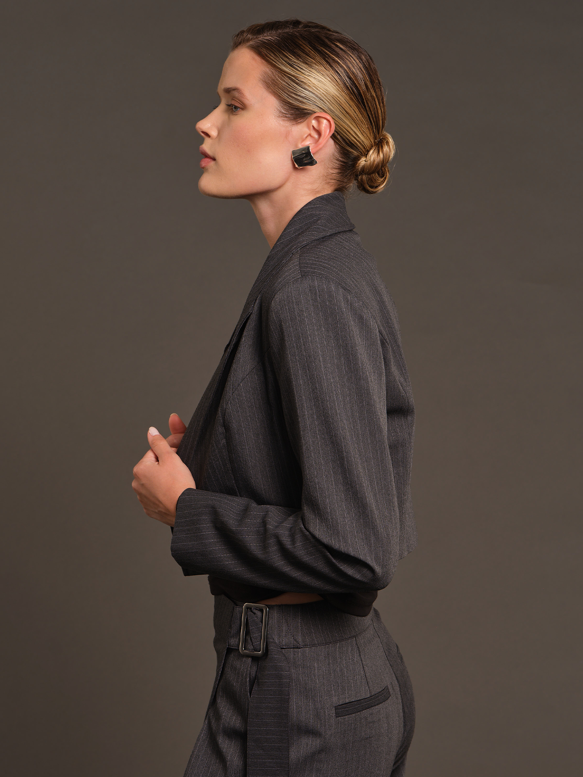 A woman in side profile wearing a dark gray pinstripe blazer and matching trousers with a belt, styled with black geometric stud earrings against a solid dark gray studio background.