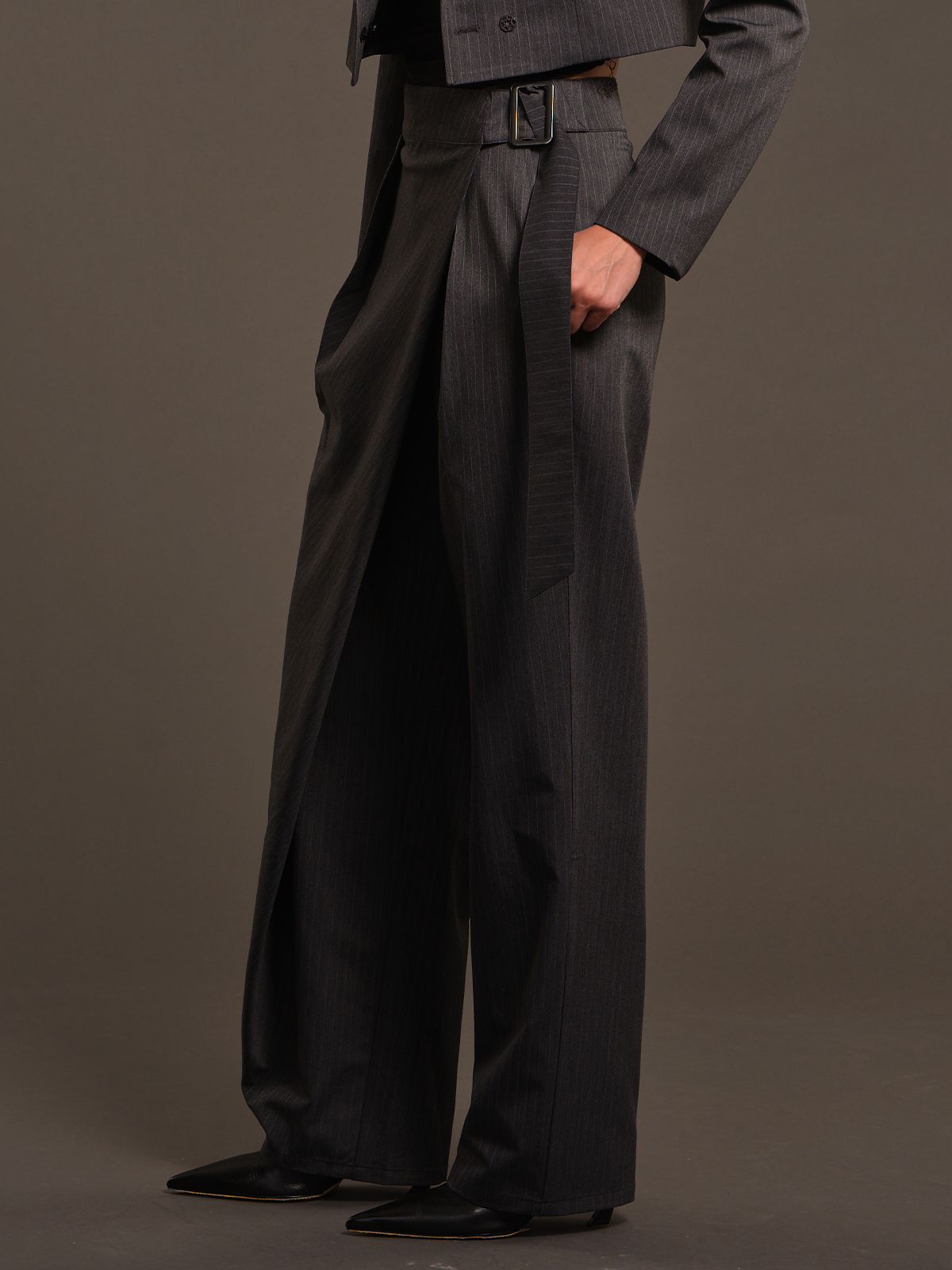 A model wearing high-waisted, wide-leg charcoal gray pinstripe trousers with a matching belt, styled with a dark pinstripe blazer and black pointed-toe shoes against a solid dark gray studio background.