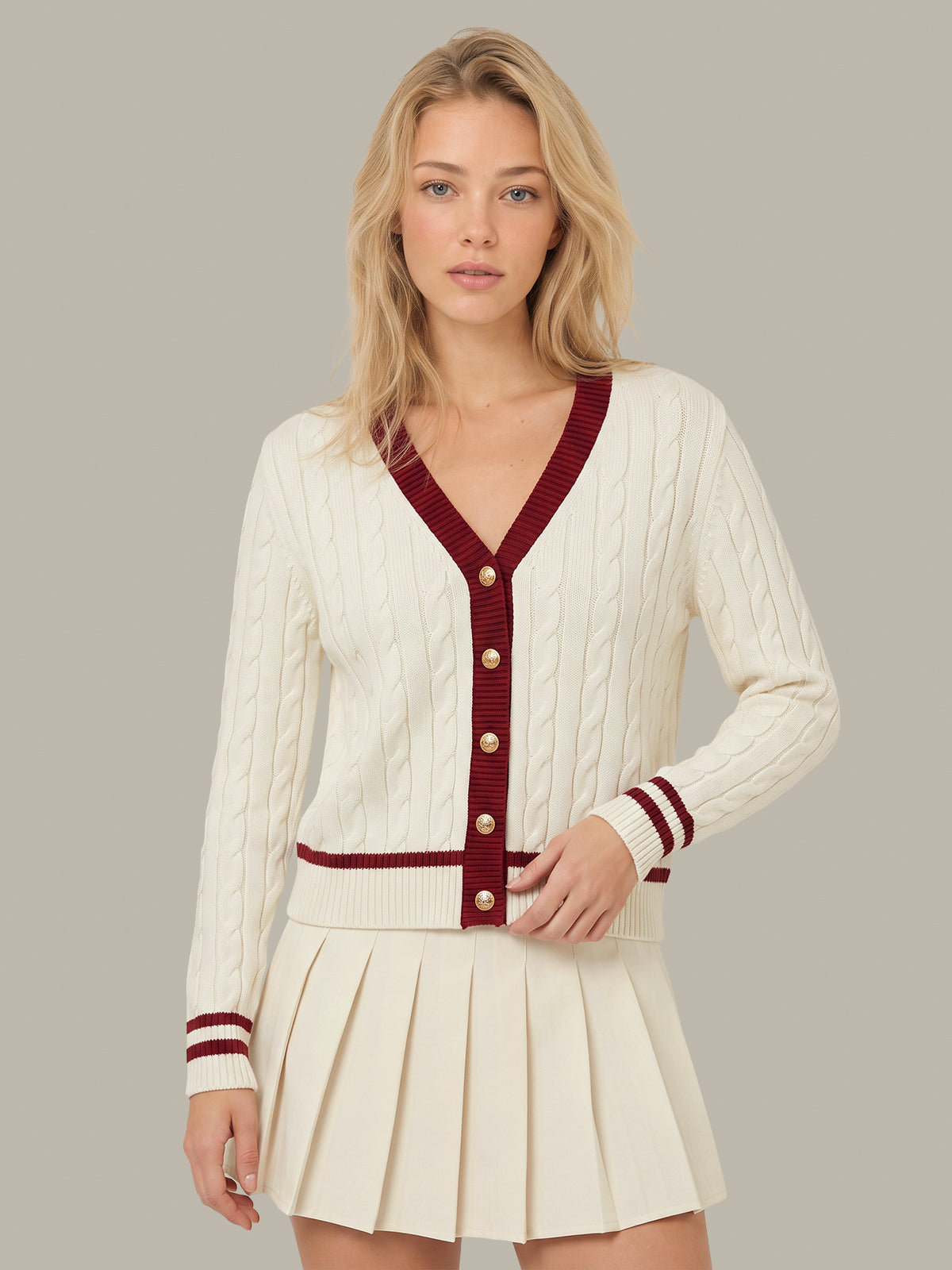 A woman modeling a cream cable-knit cardigan with burgundy trim and a matching pleated cream mini skirt against a solid taupe background.