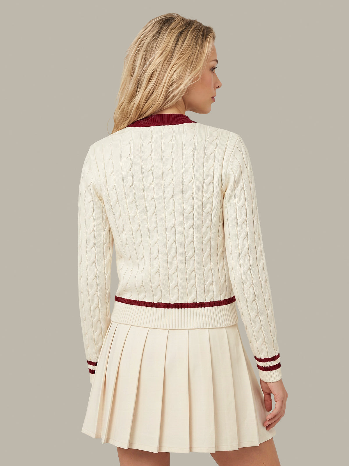 A woman shown from the back wearing a cream cable-knit cardigan with burgundy trim and a matching pleated cream skirt, against a solid taupe background.