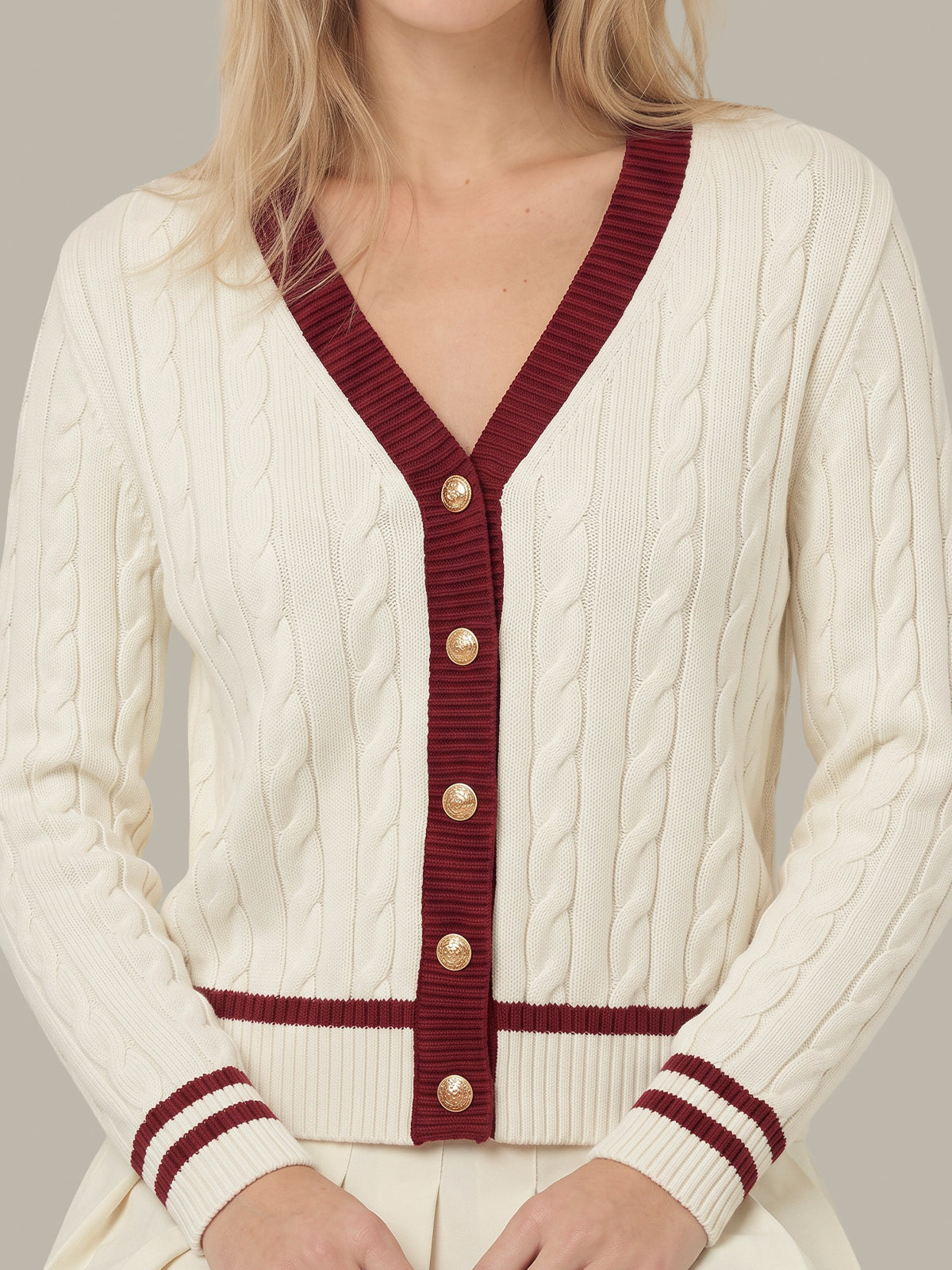 A close-up of a woman wearing a cream cable-knit cardigan with burgundy ribbed trim and gold buttons.
