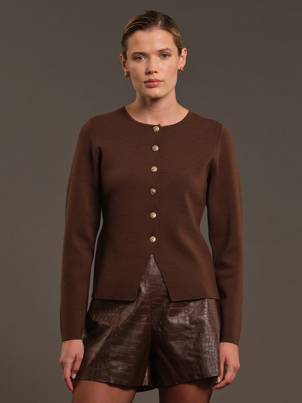 A model wearing a brown long-sleeve knit cardigan with a button-down front and brown faux-leather shorts, posed against a solid dark gray studio background.
