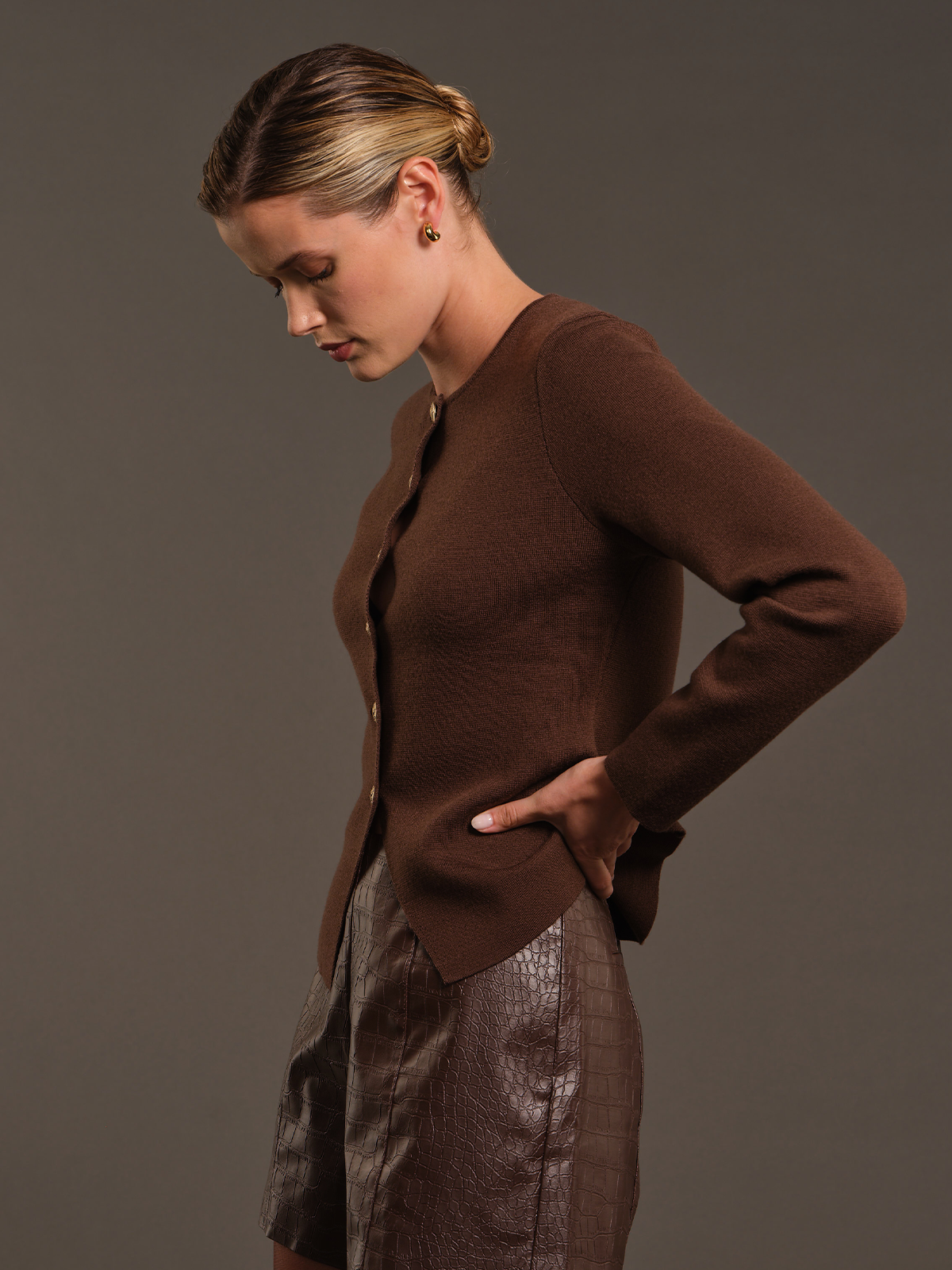 A woman wearing a brown ribbed knit cardigan and a brown textured skirt, posing in profile against a solid dark gray studio background.