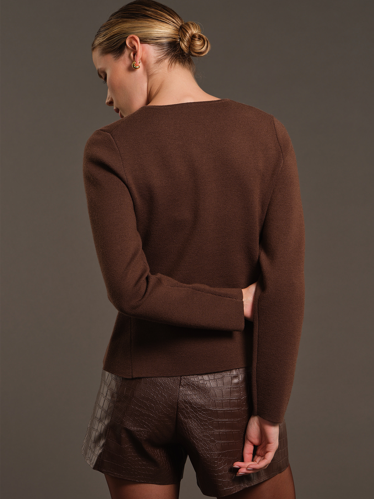 A woman shown from the back wearing a brown knit long-sleeve sweater and textured brown shorts against a plain dark gray studio background.