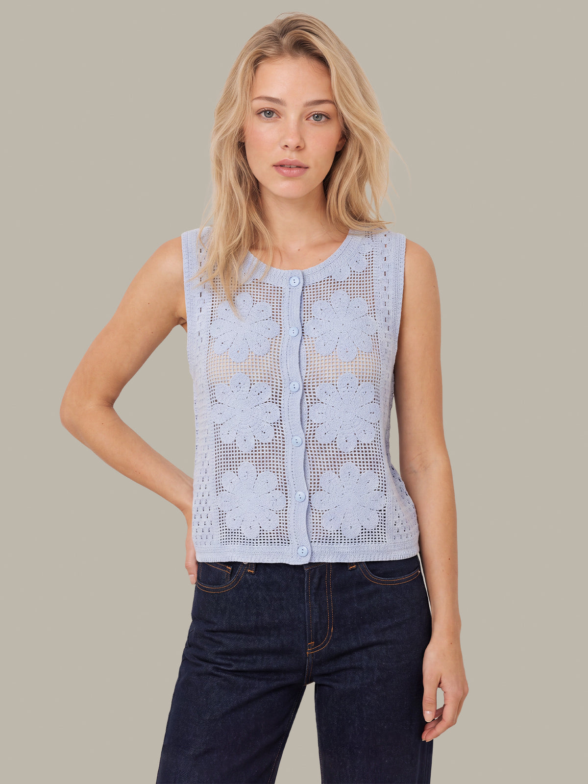 A woman modeling a sleeveless light blue crochet knit top with large floral lace patterns and a button-up front, paired with dark blue jeans, against a solid beige background.