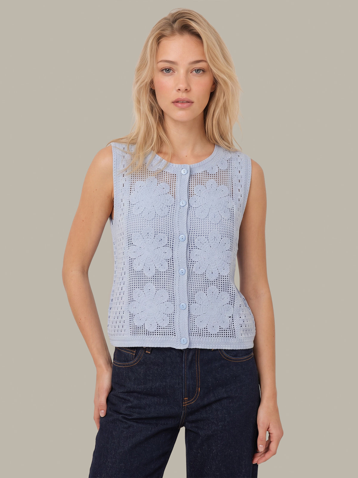 A blonde woman modeling a sleeveless light blue knit button-front top with floral lace panels, paired with dark blue jeans, against a solid taupe background.