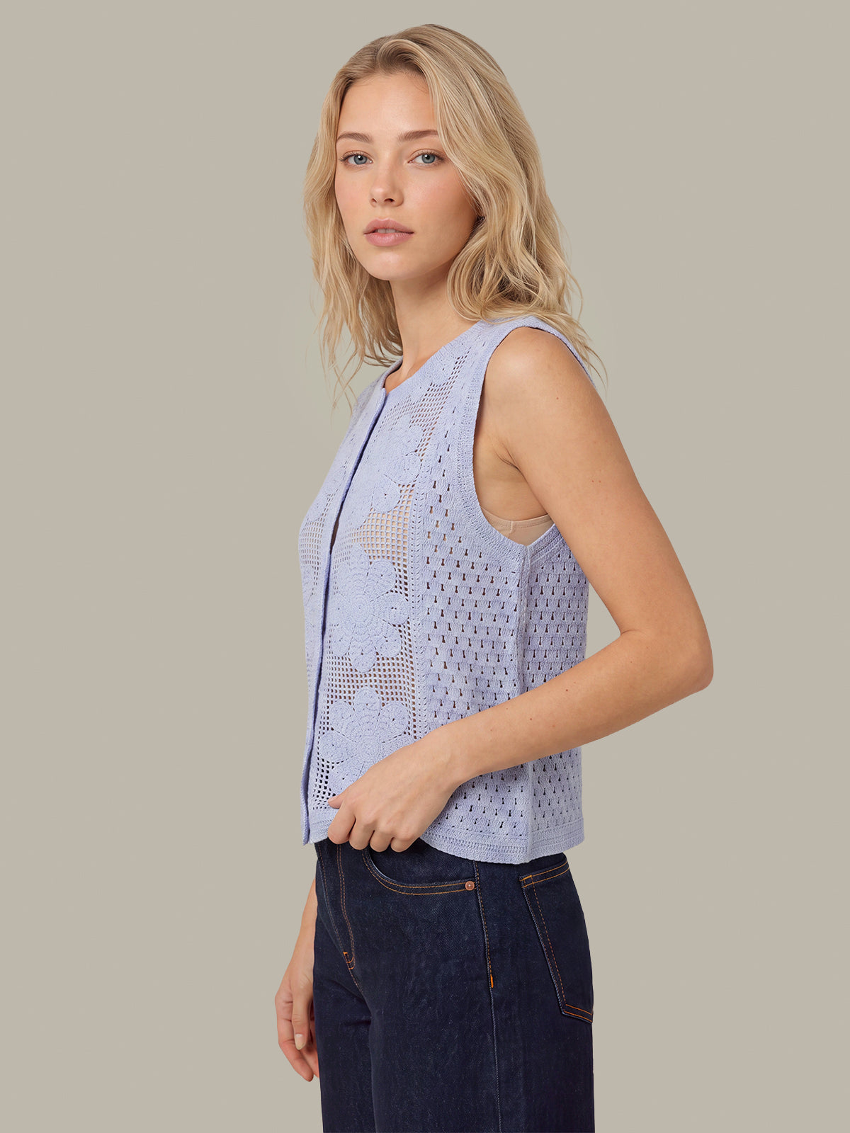 A woman wearing a light blue sleeveless crochet vest over a white top and dark blue jeans, photographed against a solid beige background.