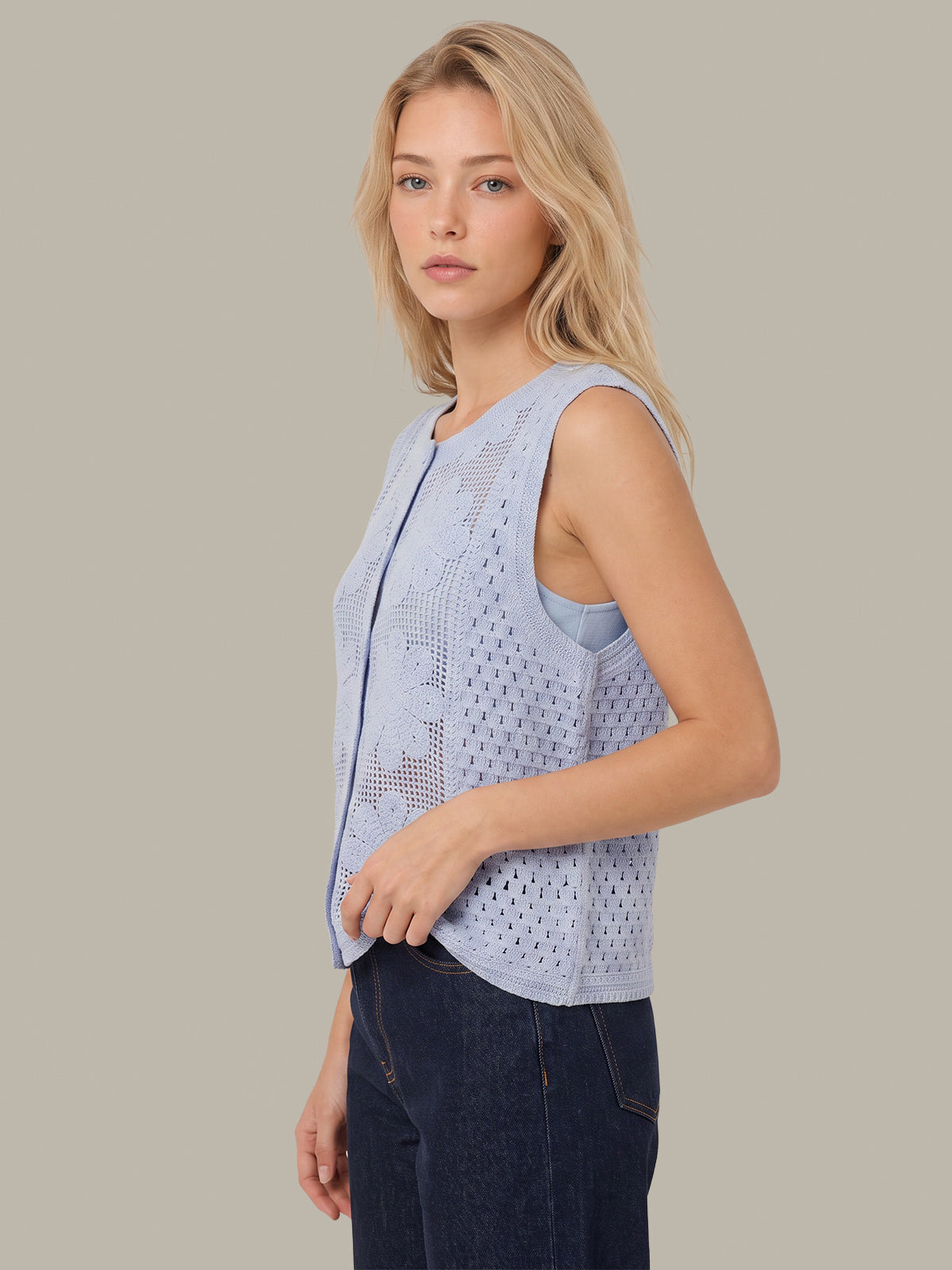 A woman wearing a light blue sleeveless knit vest with a front zipper and eyelet pattern, paired with dark blue jeans, set against a solid beige background.