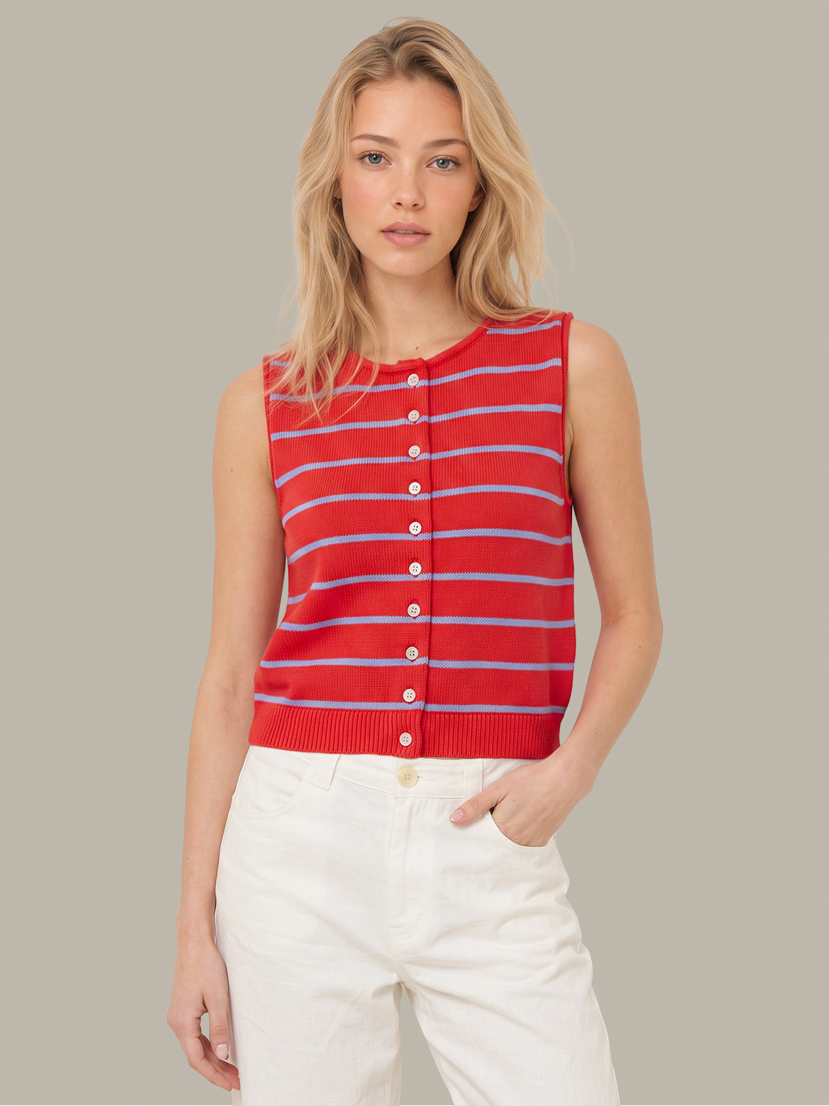 A blonde woman wearing a red sleeveless knit top with thin blue and white horizontal stripes and a button-down front, paired with white pants, against a solid gray background.