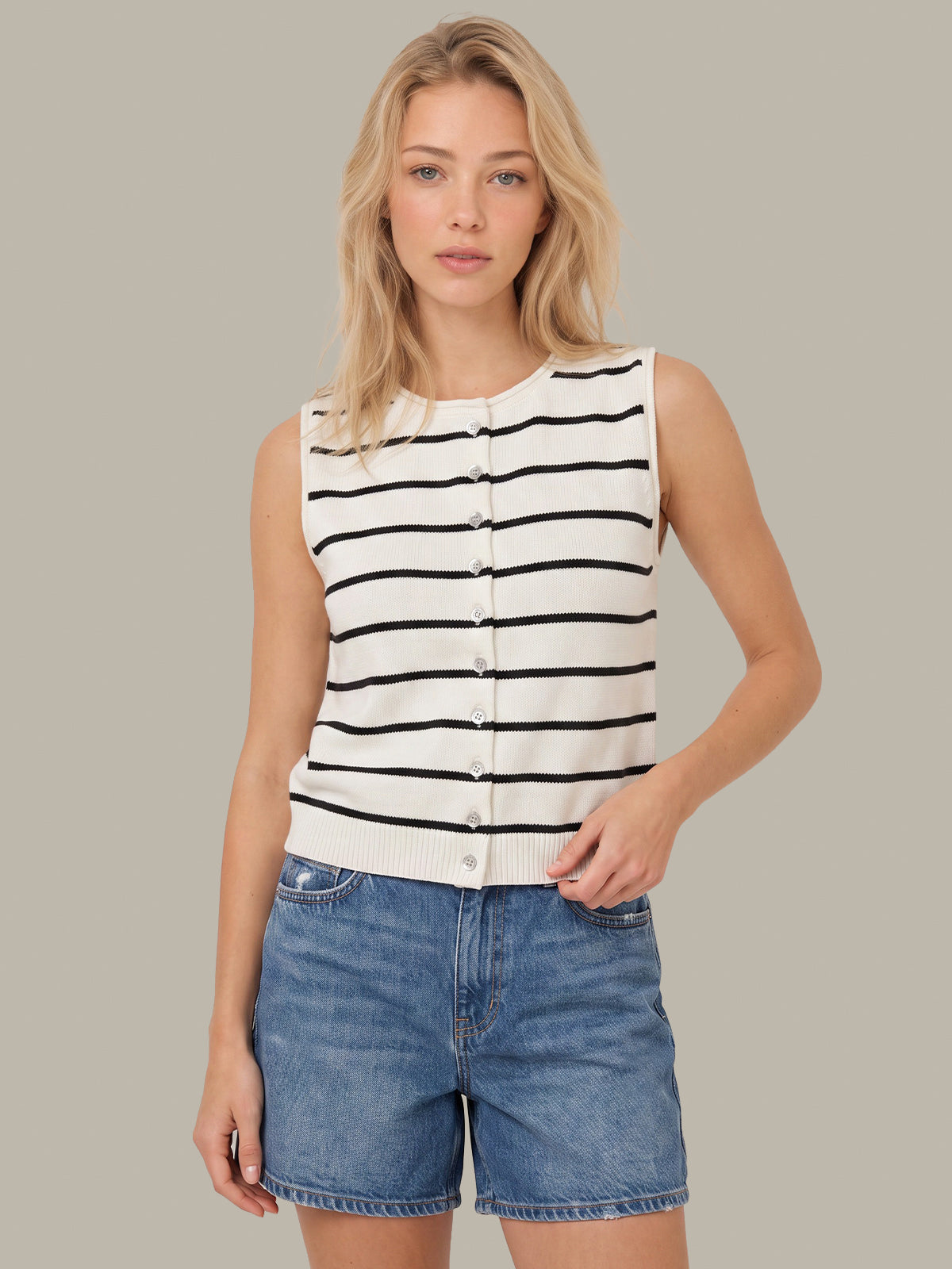 A woman modeling a sleeveless knit top with black horizontal stripes on an off-white background, featuring a button-down front. She is paired with blue denim shorts against a solid neutral gray background.