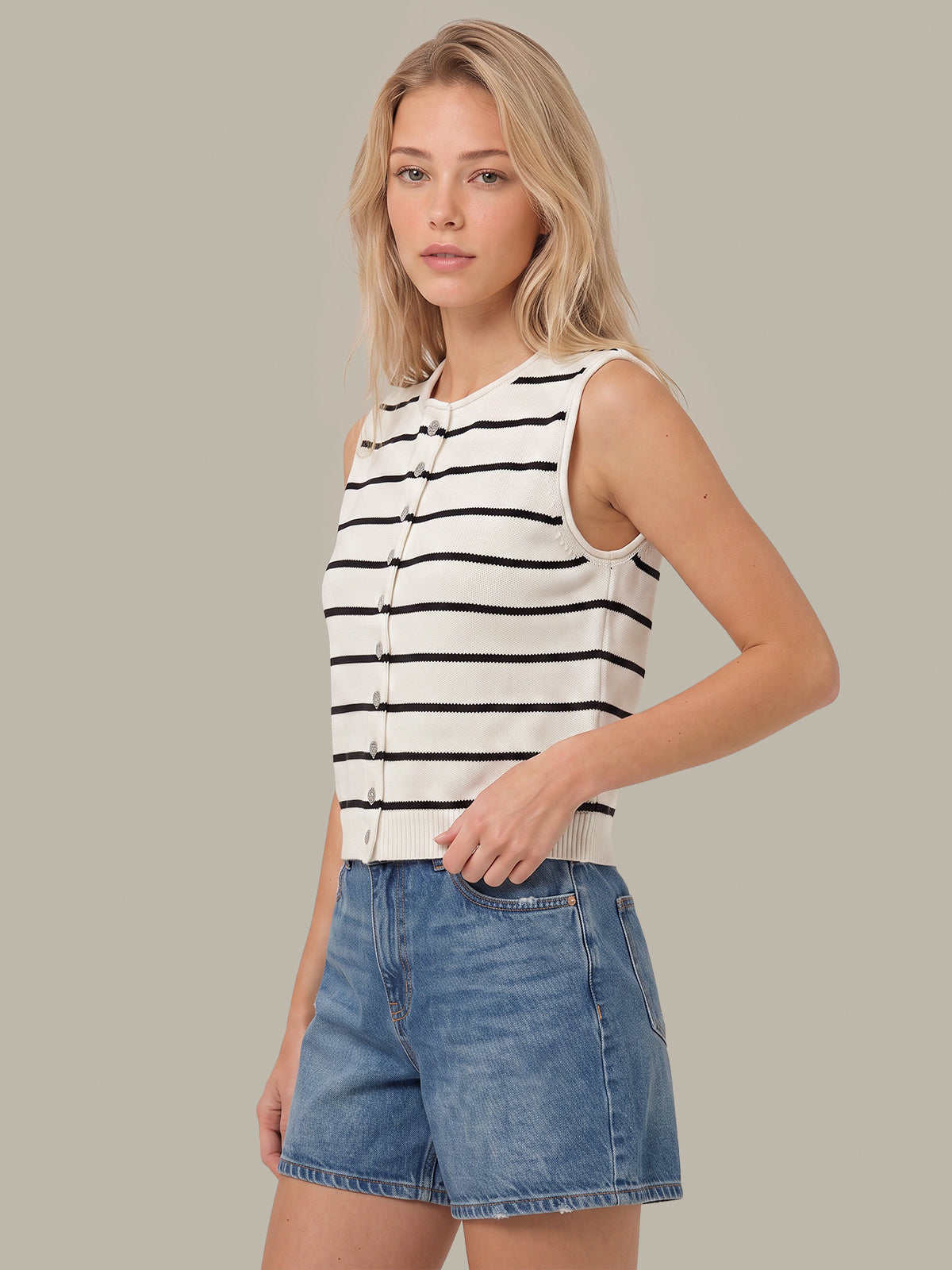 A woman modeling a sleeveless knit top with black horizontal stripes on an off-white background, paired with blue denim shorts, against a solid taupe background.
