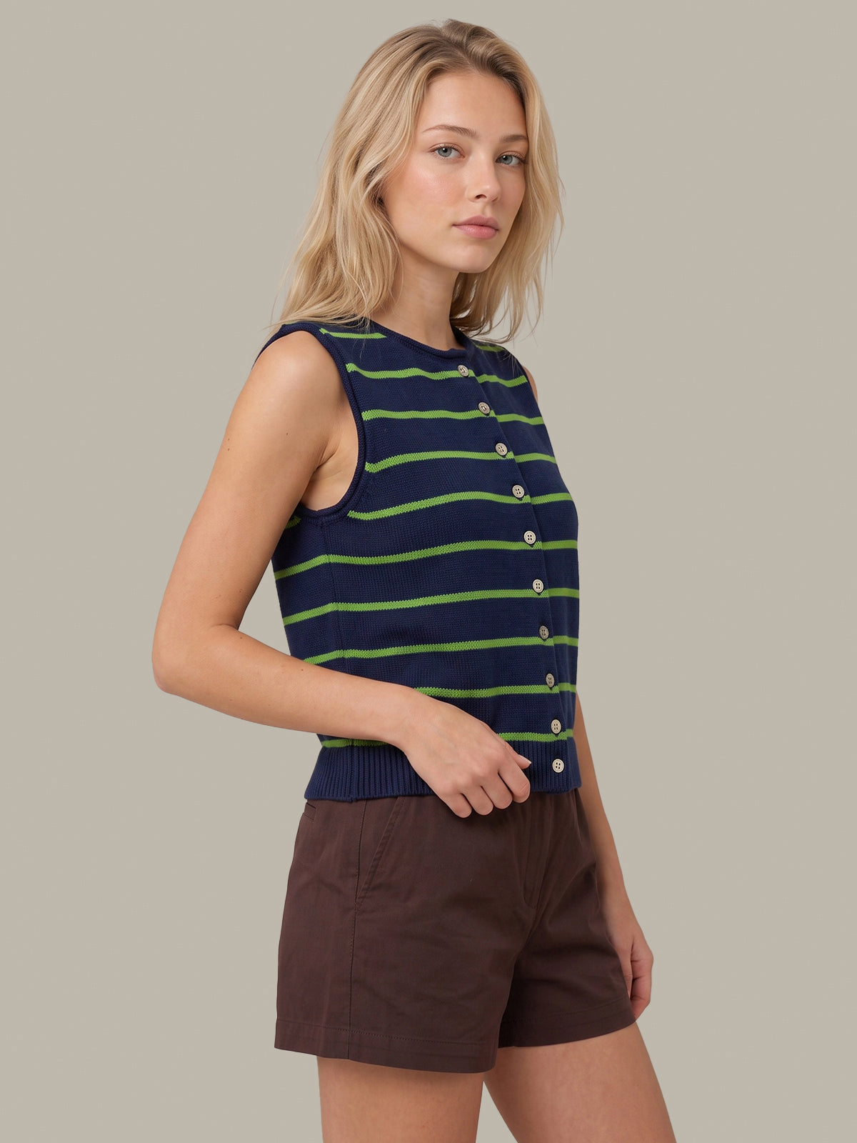 A woman modeling a sleeveless navy knit top with green horizontal stripes and a button placket, paired with brown shorts, against a solid light gray background.