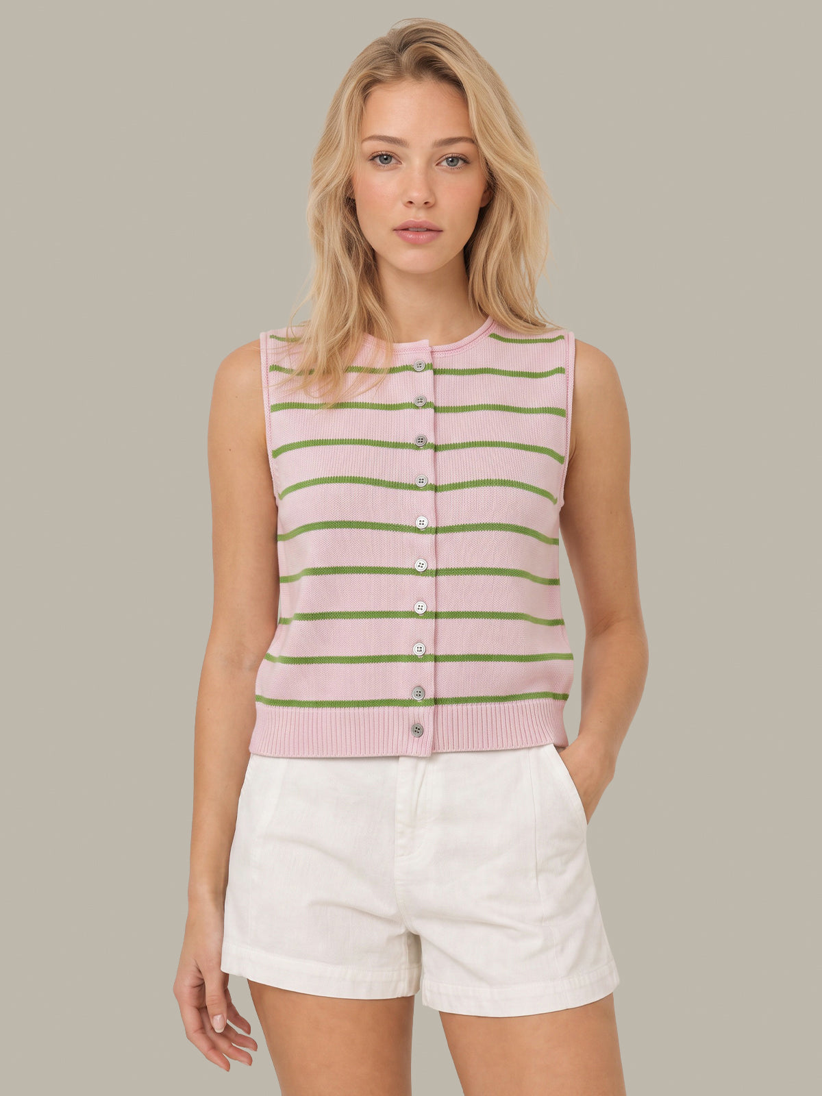 A woman modeling a sleeveless pink knit vest with thin green horizontal stripes and a button front, paired with white shorts, against a solid taupe background.