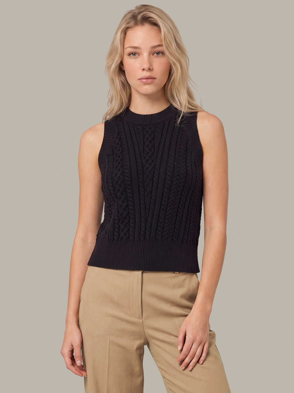 A blonde woman wearing a sleeveless black cable-knit mock-neck sweater vest paired with beige trousers, standing against a solid light gray background.